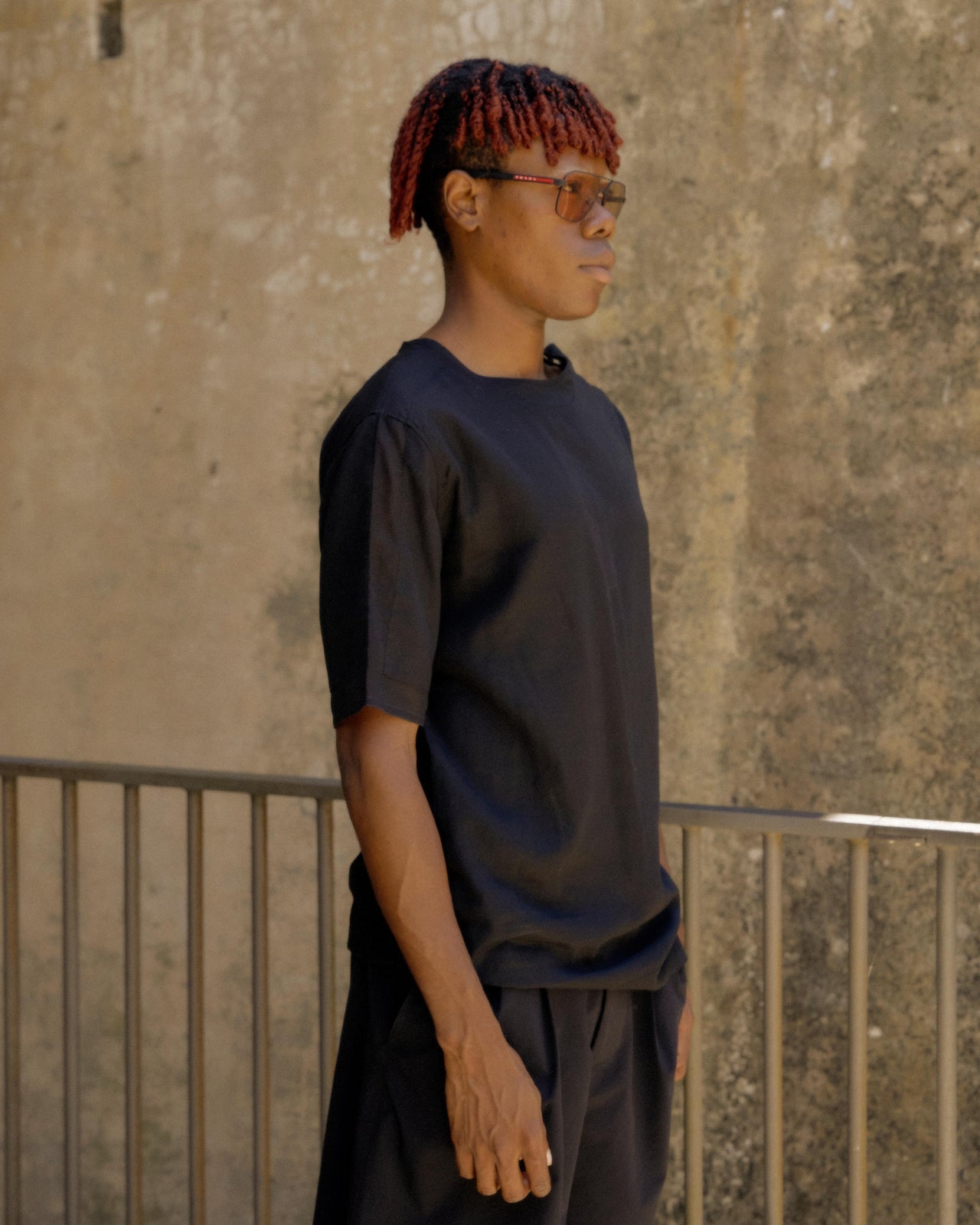 A person wearing a black sheer Hermes t-shirt with three button closures at the neck, standing in a casual pose with one hand in a pocket.