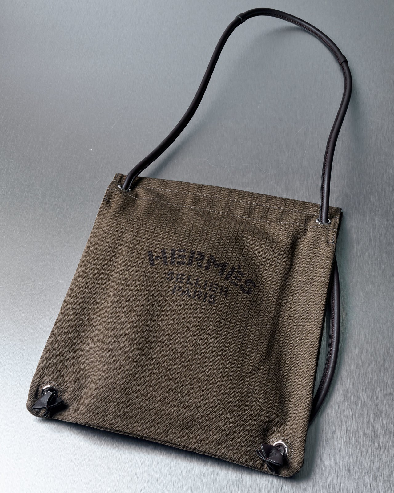 Olive green Hermes bag with black text "Hermes Sellier Paris" on the front, featuring black rope handles and a minimalist design on a metallic surface.