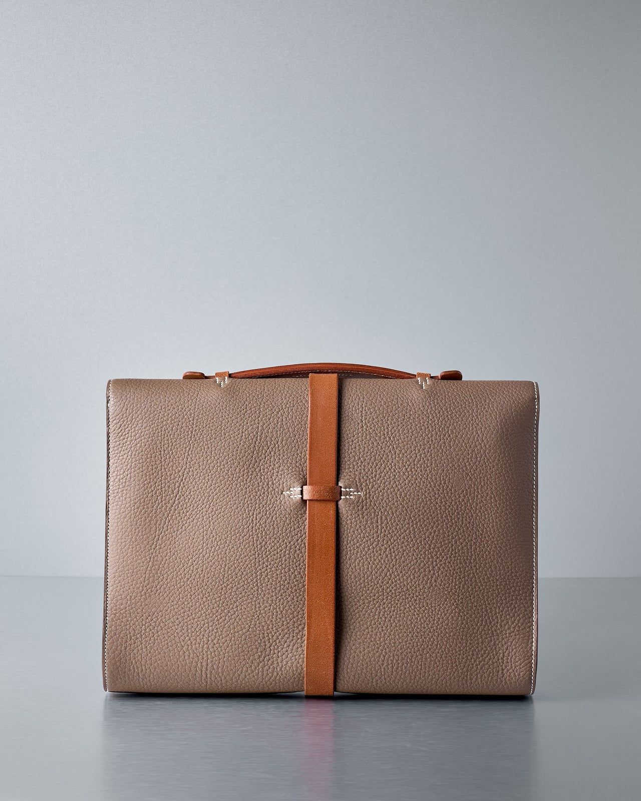 Brown Hermès leather briefcase with a brown strap on a grey background.
