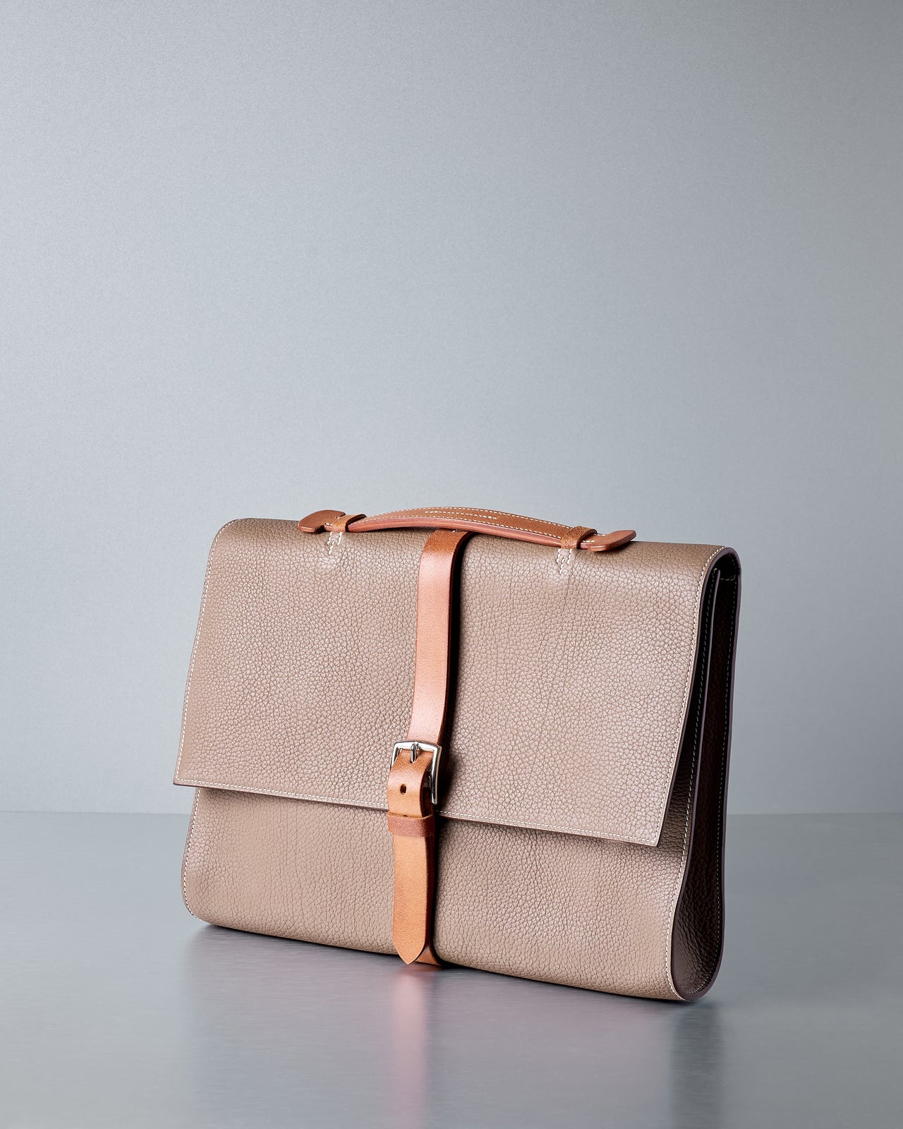 Brown Hermès leather briefcase with a brown strap on a grey background.