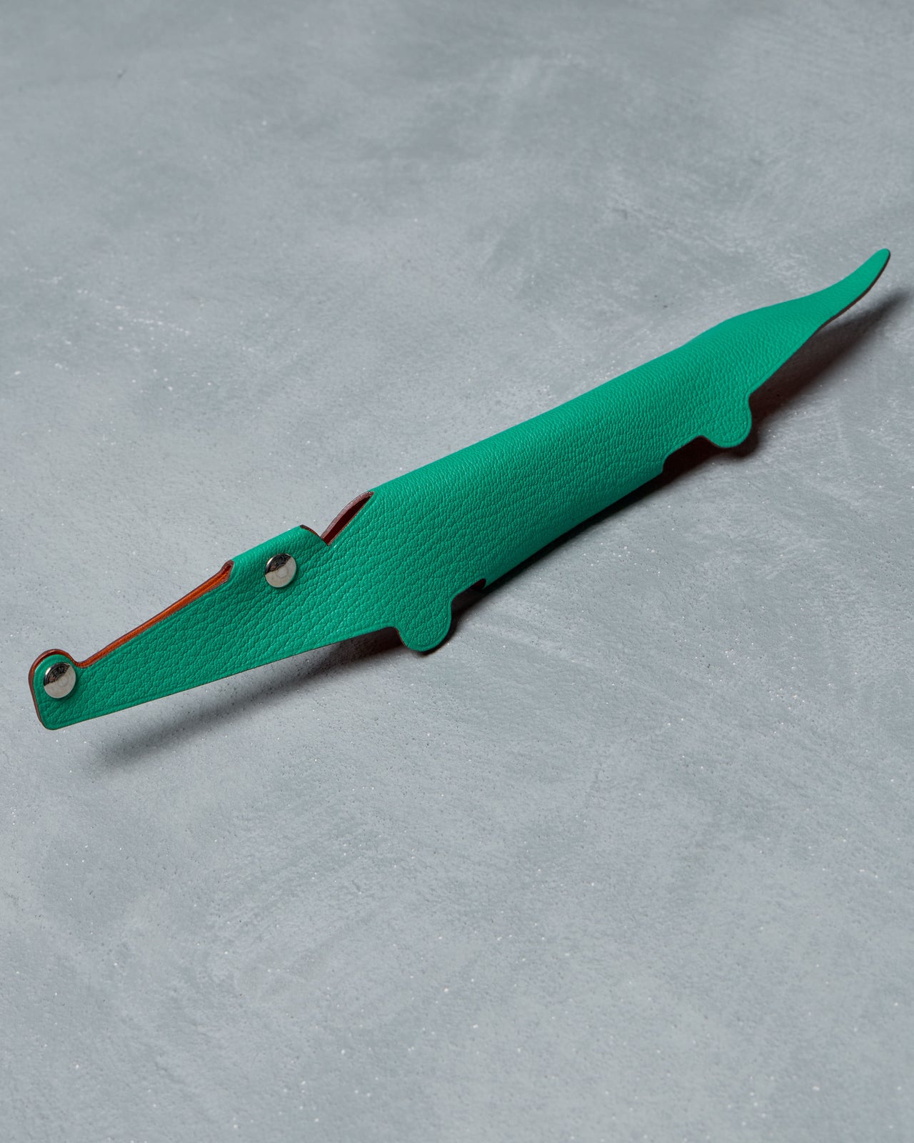 Green crocodile-shaped art object from Hermès with silver rivets, set against a textured gray background. Sleek and playful design, exuding a whimsical tone.