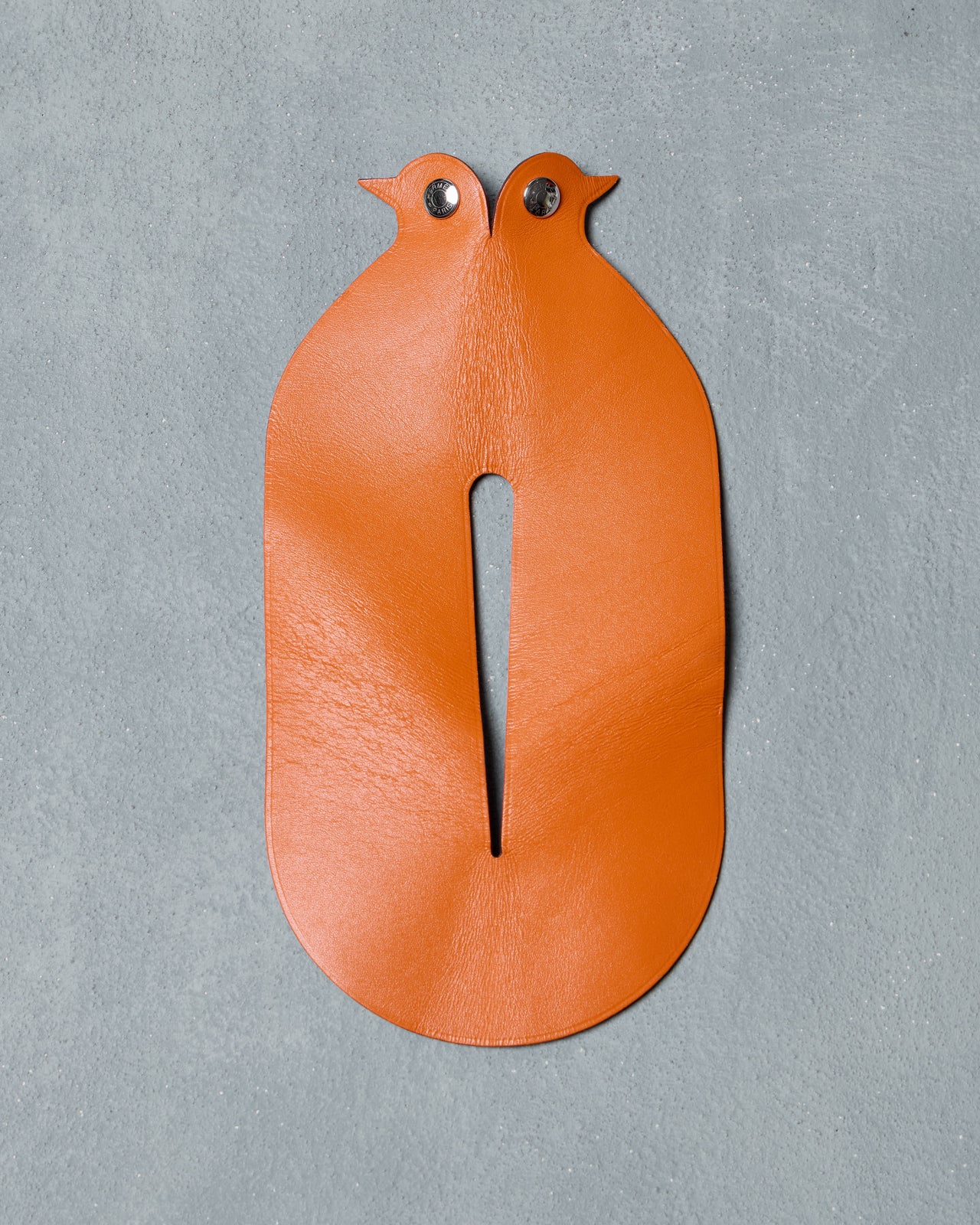 A minimalist, abstract bird sculpture from Hermès made from orange and blue leather, featuring a shiny metal eye, on a textured gray surface. Modern and playful design.