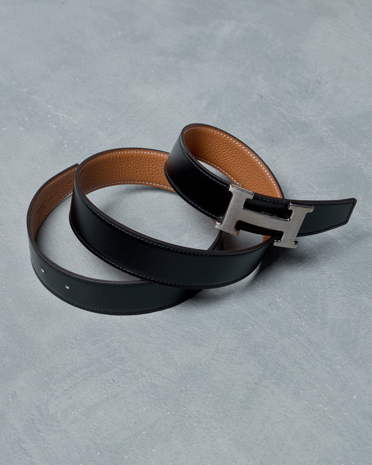 Black leather Hermes belt with a silver buckle on a gray surface