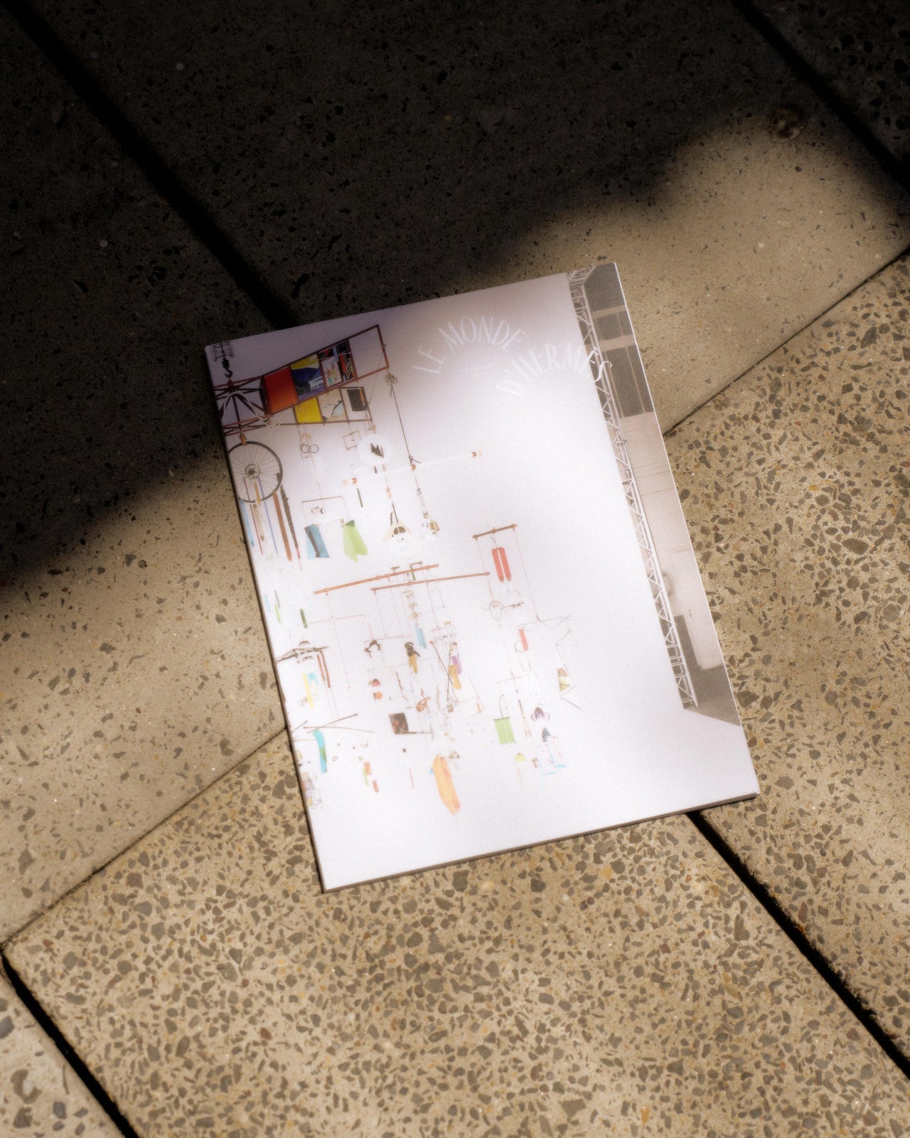 A magazine from Hermès titled "Le Monde" lies on a sunlit, textured pavement. The cover features abstract art in bright colors. The scene feels casual and serene.