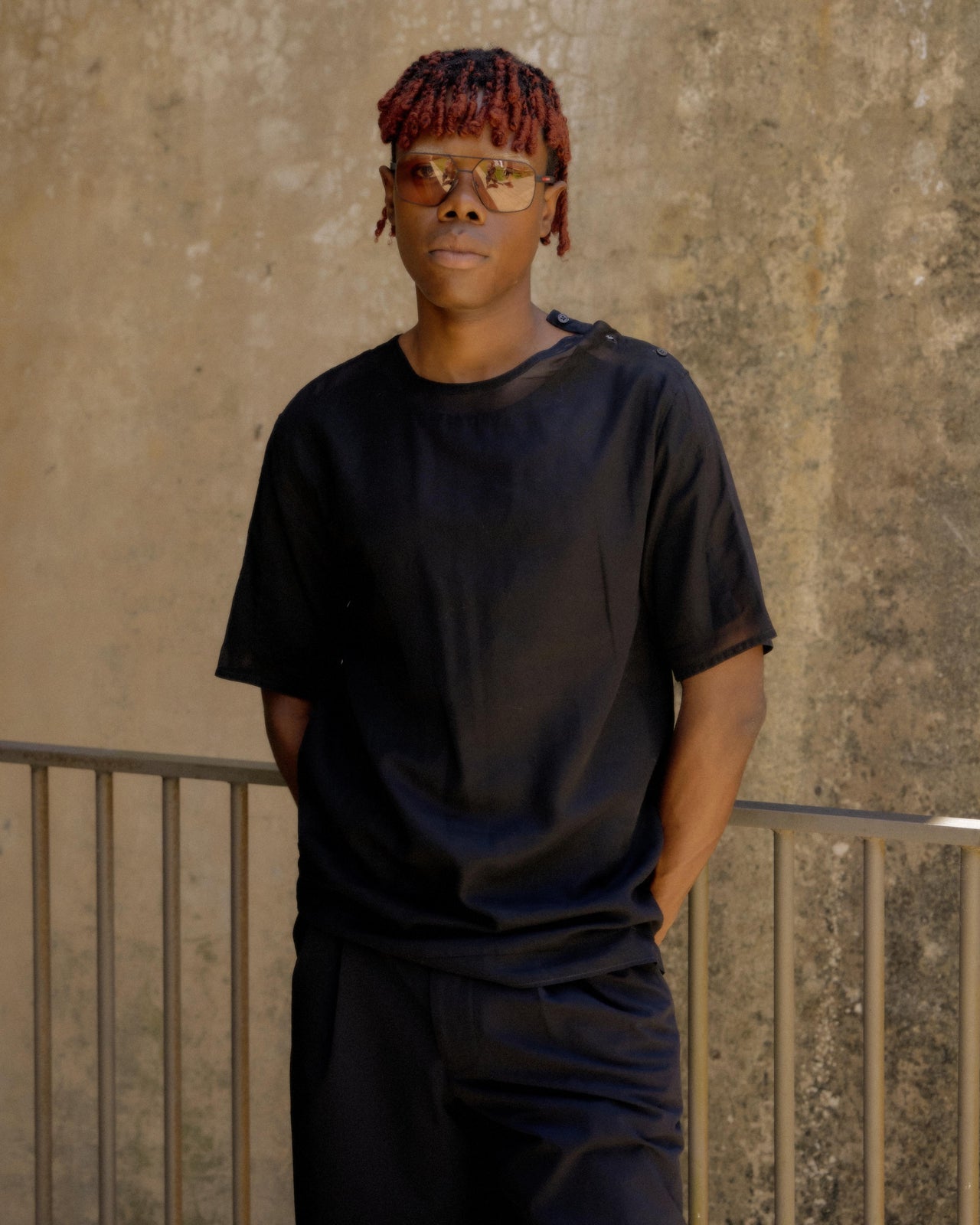 A person wearing a black sheer Hermes t-shirt with three button closures at the neck, standing in a casual pose with one hand in a pocket.
