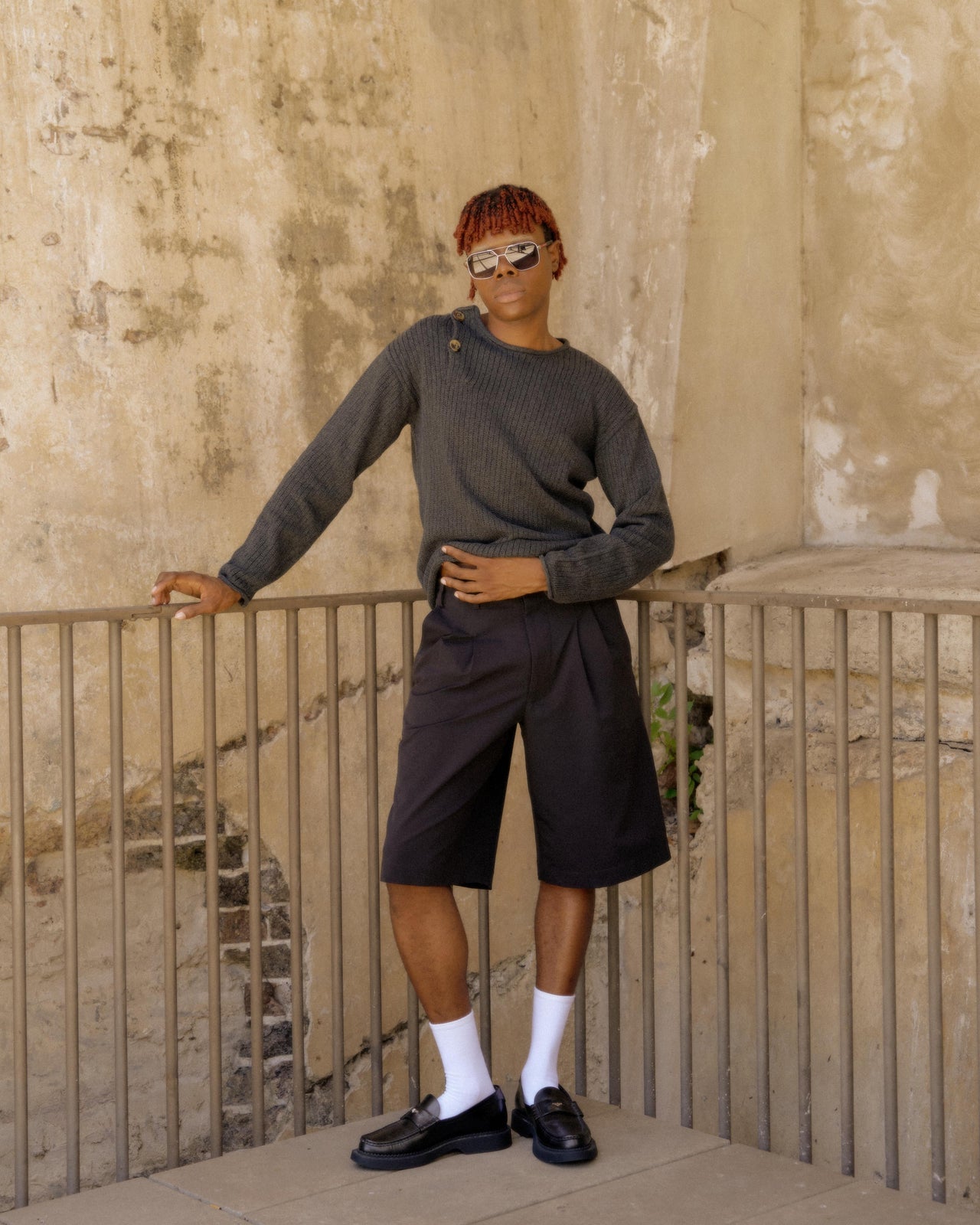 A person standing with one hand on a rail, wearing a grey knitted Hermès pullover and black shorts, sunglasses, and white socks.