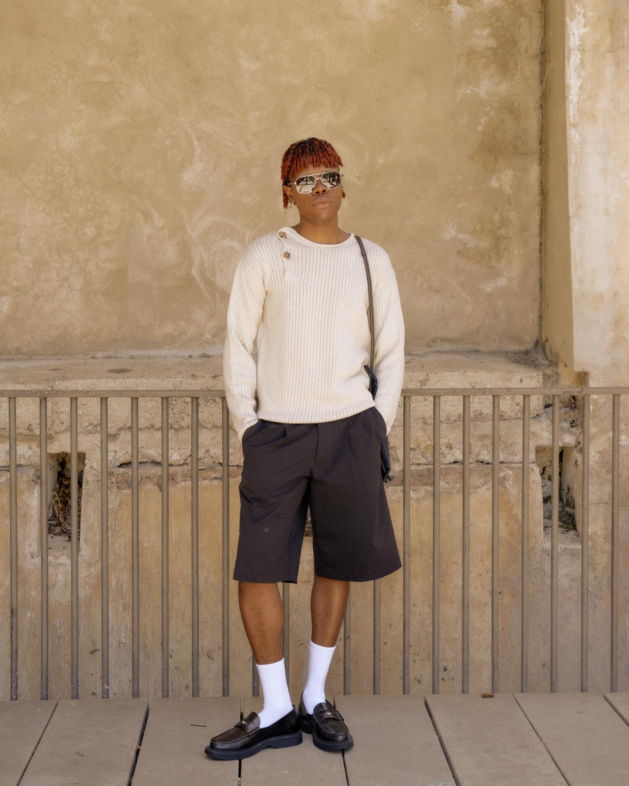 A person standing with one hand in a pocket, wearing a beige knitted Hermès pullover and black shorts, against a textured background.