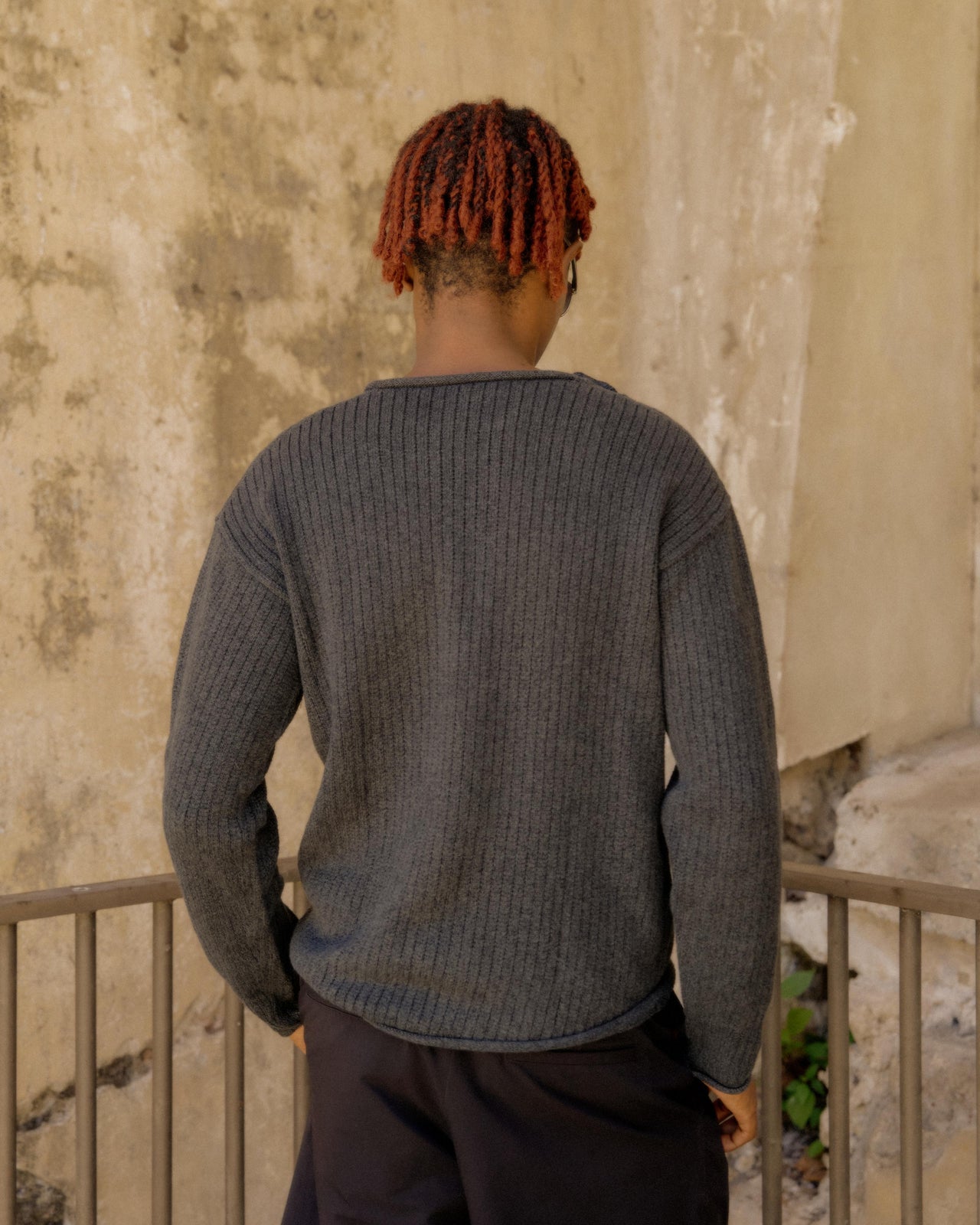 A person standing with one hand on a rail, wearing a grey knitted Hermès pullover and black shorts, sunglasses, and white socks.