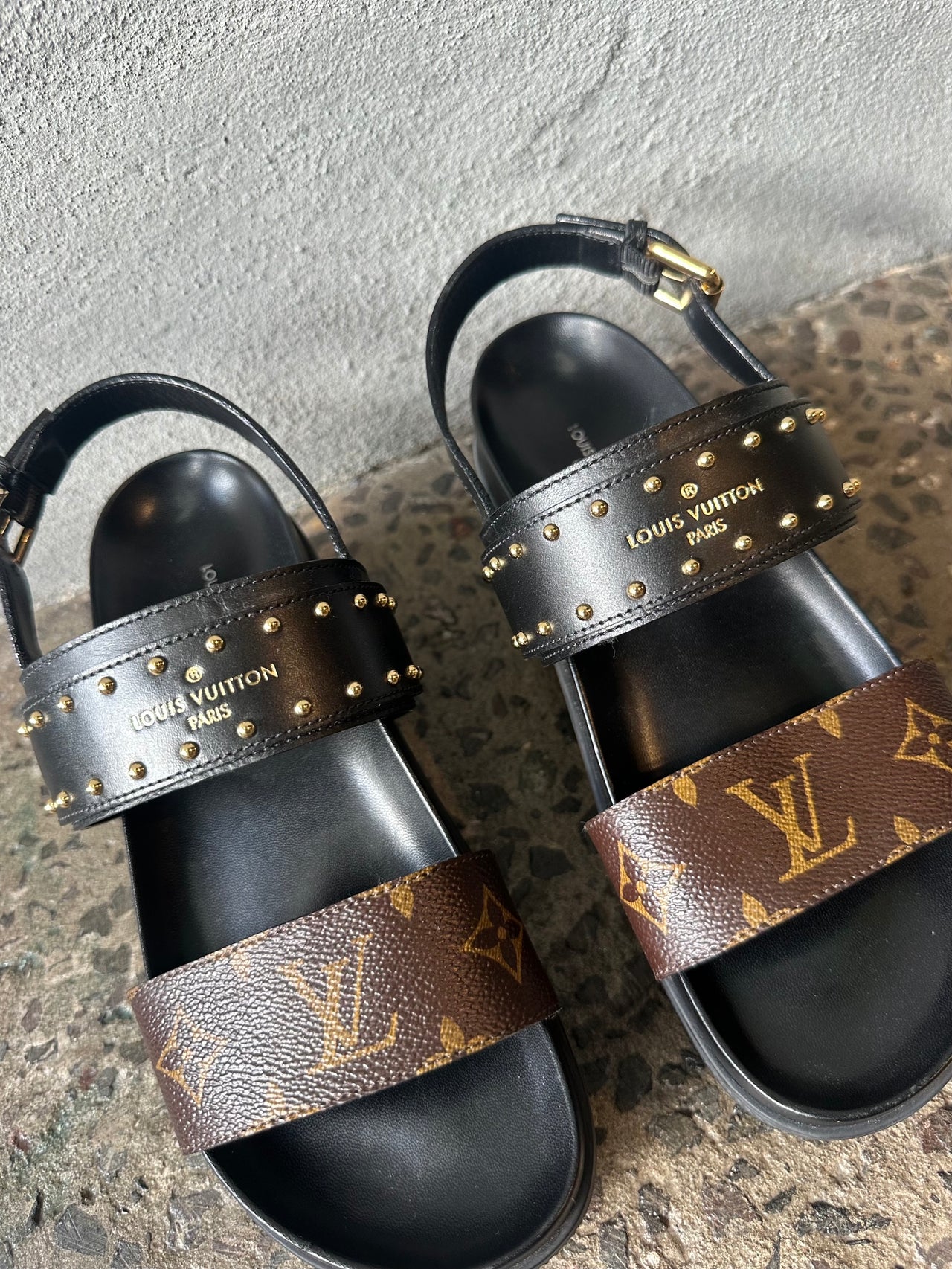 Black sandals with brown strap on a textured surface against a gray wall