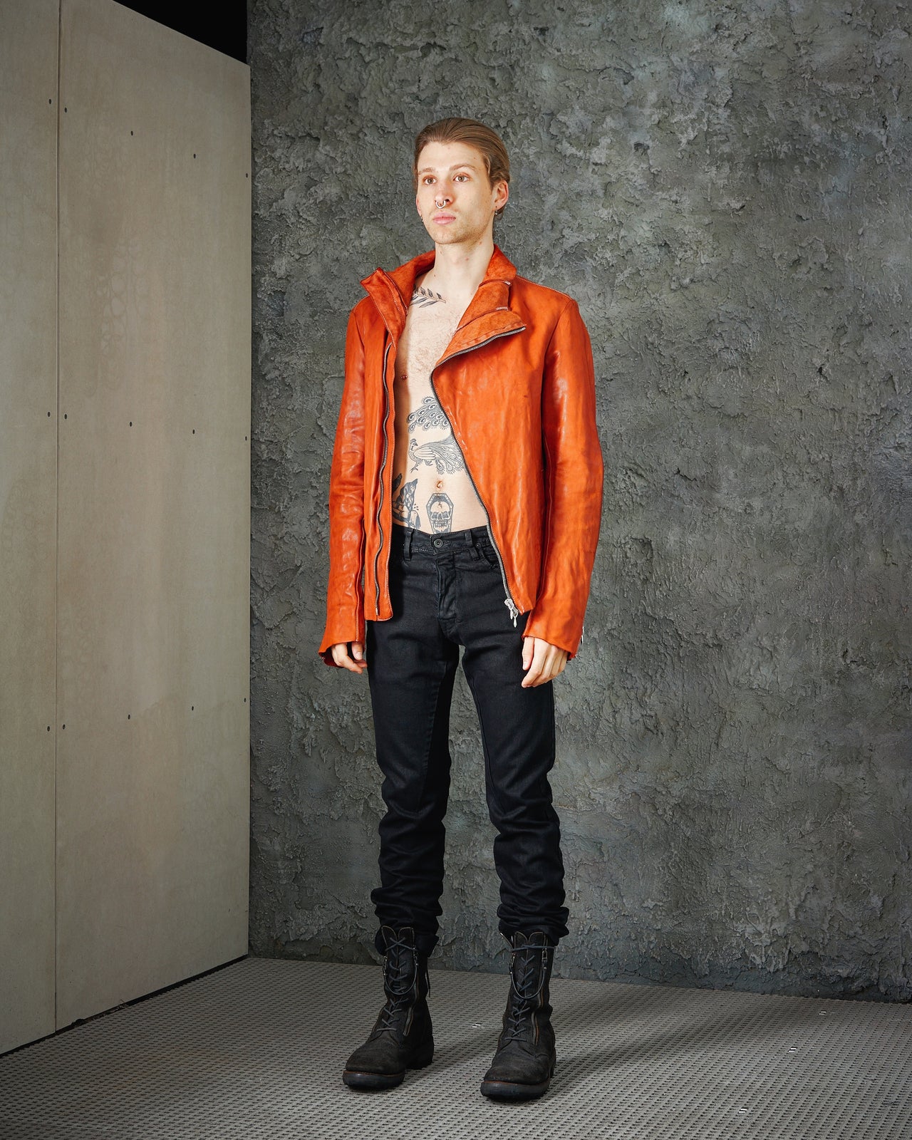 A male model wearing an orange, asymmetrical Incarnation zip leather jacket with a sculptural collar and darted structure, paired with black pants and black boots.