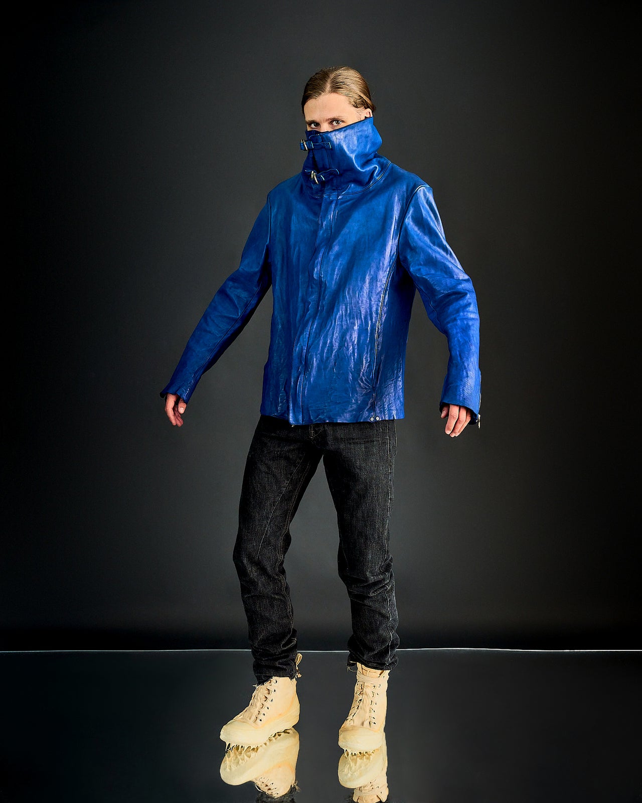 A person wears a shiny blue Incarnation jacket with a high collar covering their mouth, black pants, and tan boots