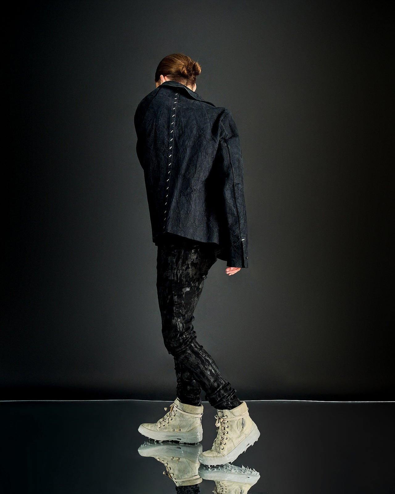 A person standing in a dark setting, wearing a black leather Isaac Sellam jacket with a front zip and textural details, paired with black pants. The person has tattoos and is accessorised with a beige hat and shoes.