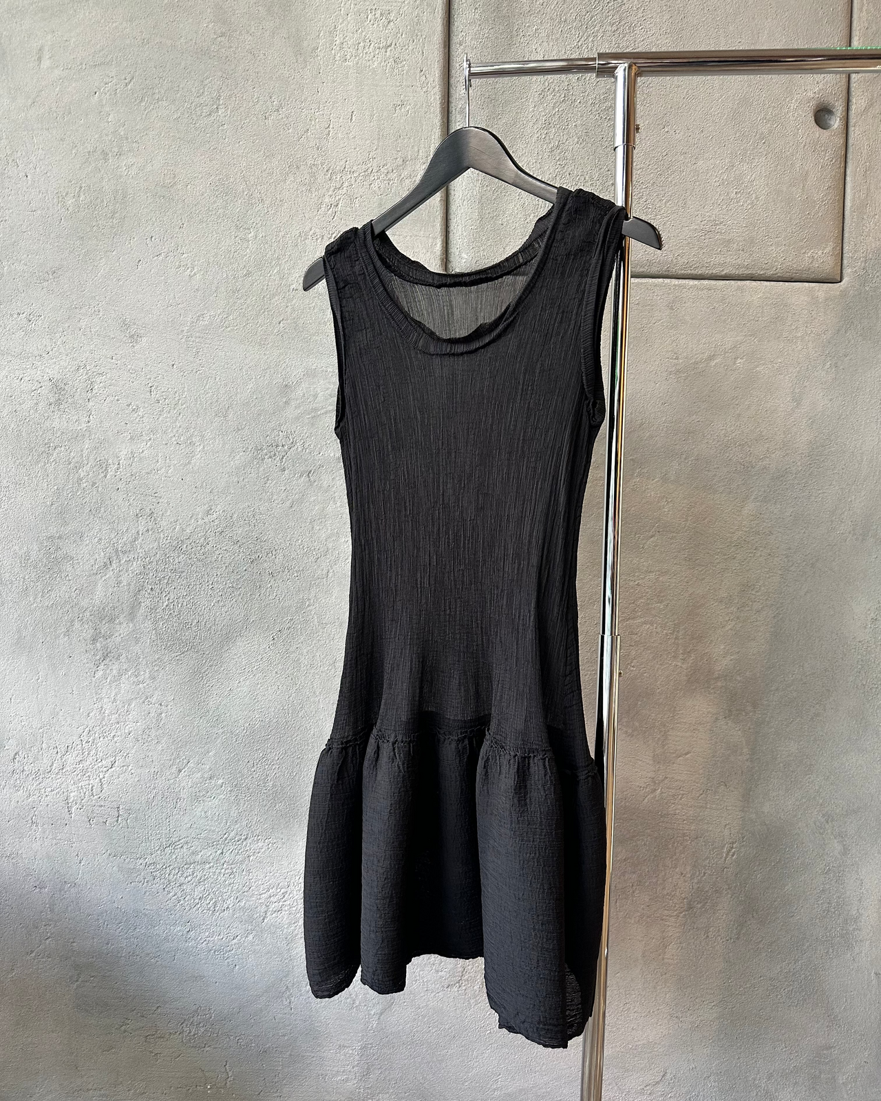 Black sleeveless Issey Miyake dress hanging on a rack against a textured wall.