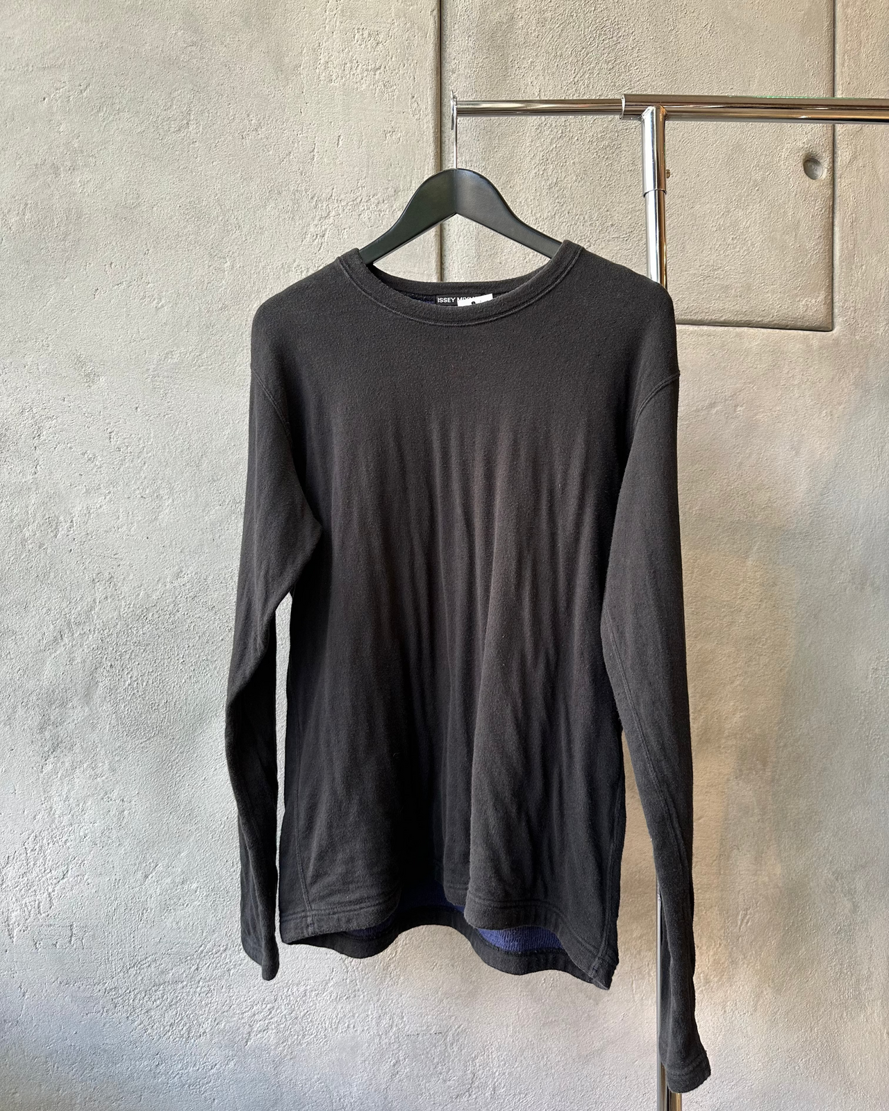 Black long-sleeve Issey Miyake sweater on a hanger against a textured gray wall