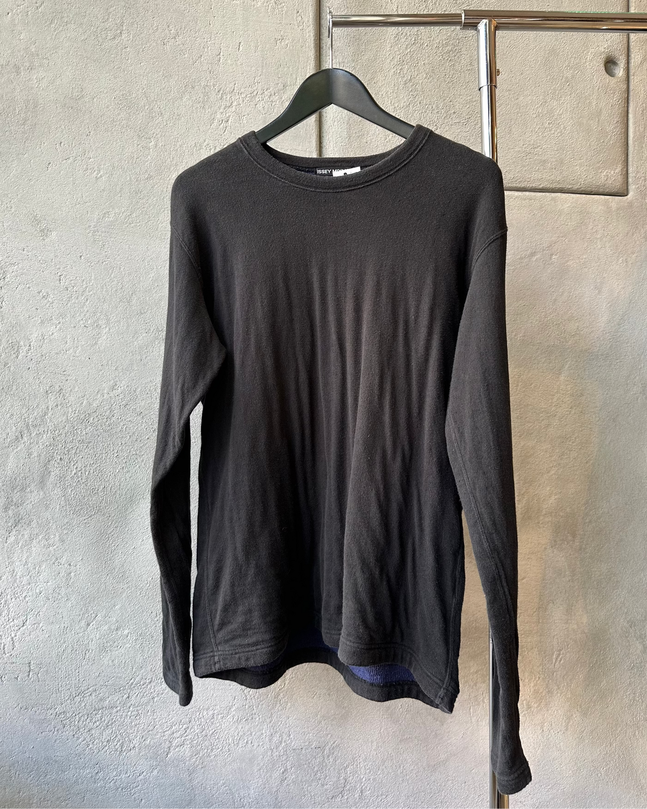 Black long-sleeve Issey Miyake sweater on a hanger against a textured gray wall