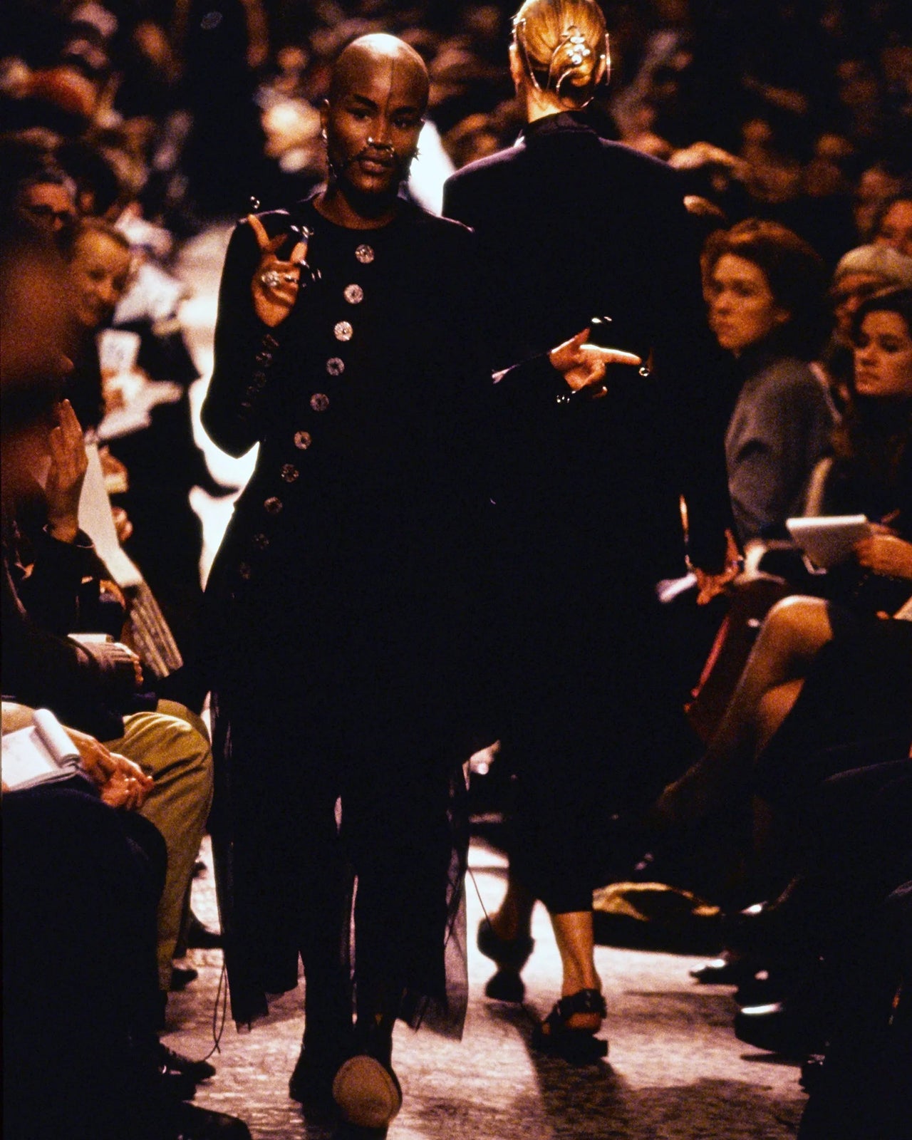 Two models walking down a Jean Paul Gaultier runway at a fashion show with an audience on either side.