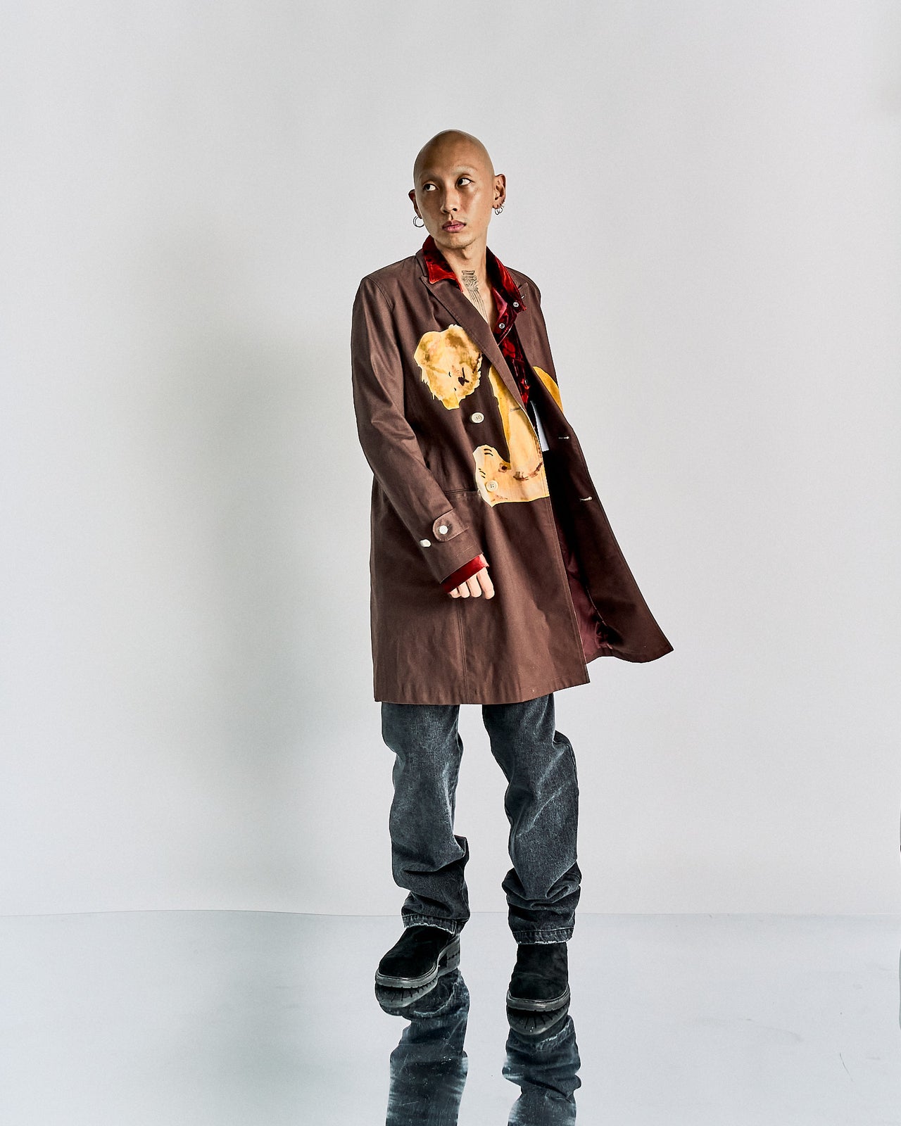 A person standing against a white background, wearing a double-breasted John Undercover chester coat with a graphic of a headless teddy bear on the front, paired with grey trousers and black boots.