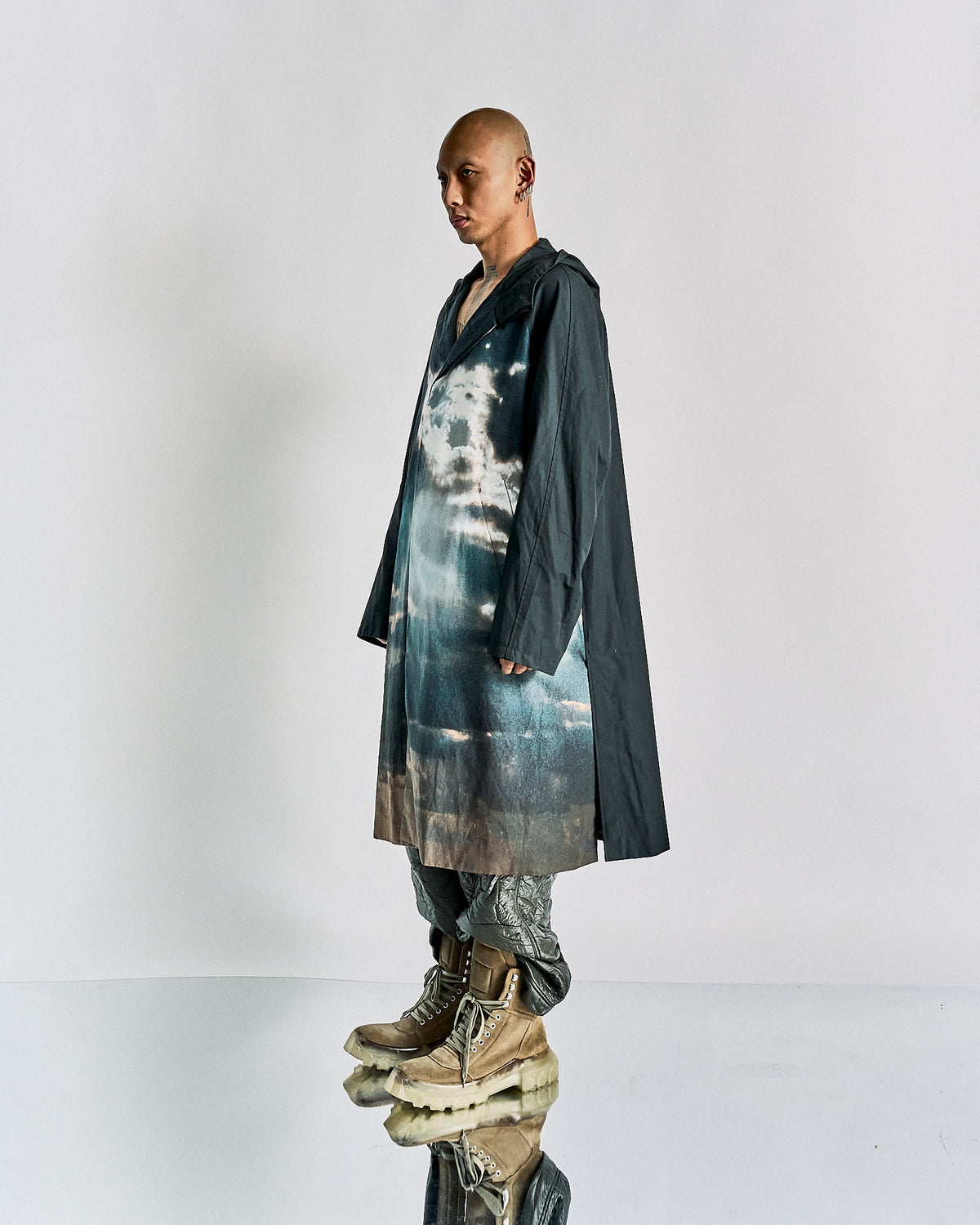A person standing wearing a black John Undercover hooded coat with a landscape panel featuring a cloud and sky pattern. The coat has a button-up front and hip pockets.