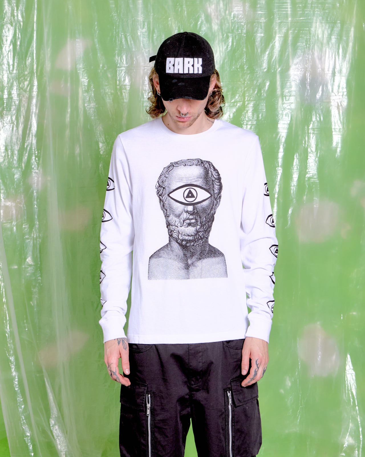 Person wearing a black cap with "BARK," a white shirt from Japanese brand Undercover, featuring a surreal face with an eye motif, and black pants. Background is green plastic. Urban and edgy vibe.