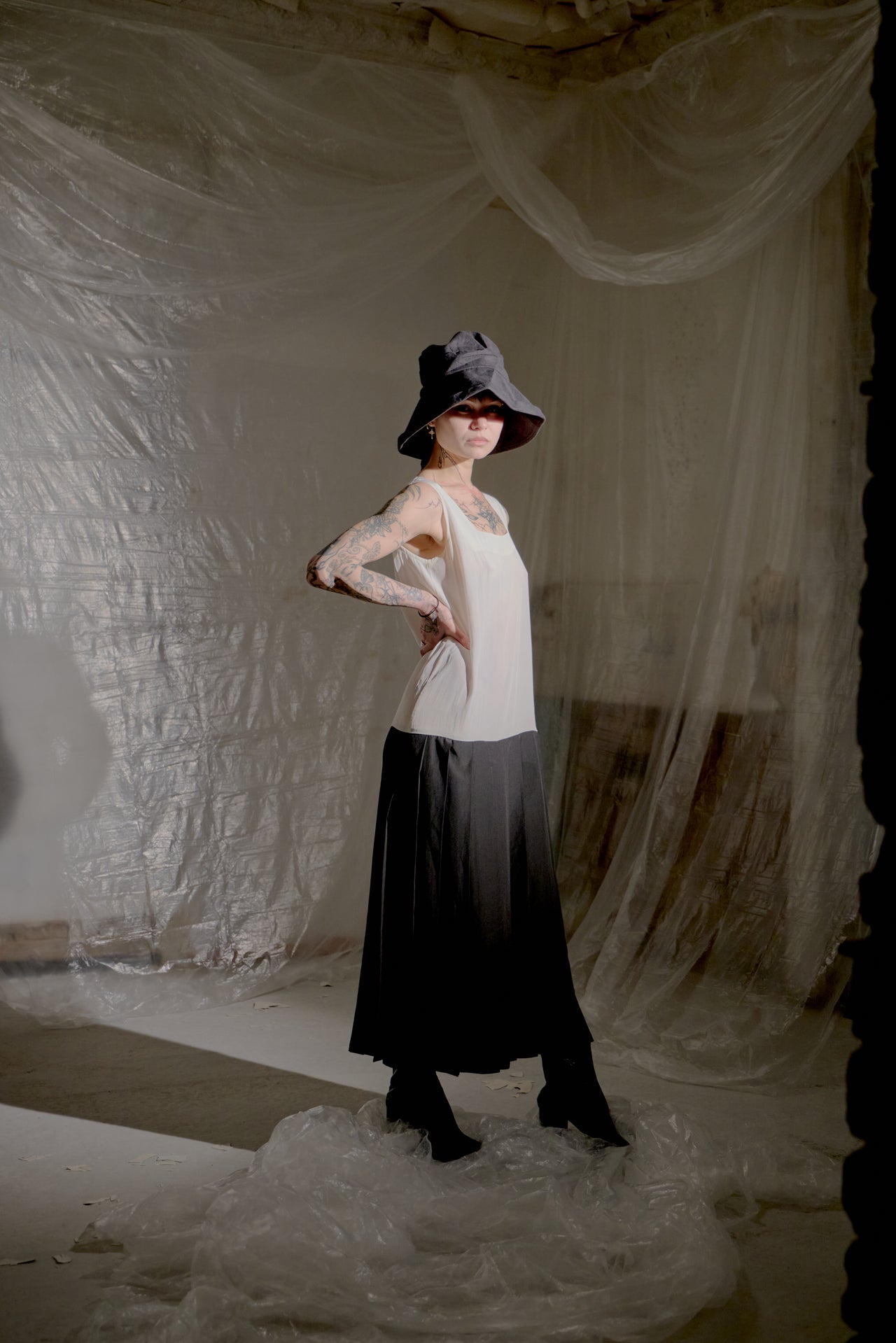 Person wearing a black and white Limi Feu dress with a hat, standing in a softly lit room with draped fabric.