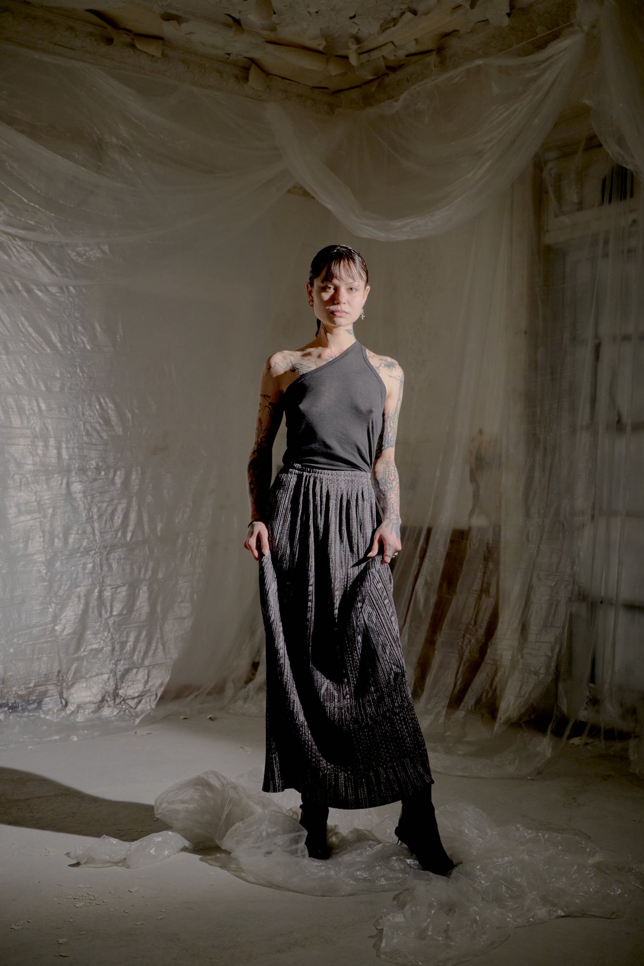 Person wearing a dark top and skirt standing in a textured, neutral-coloured room.