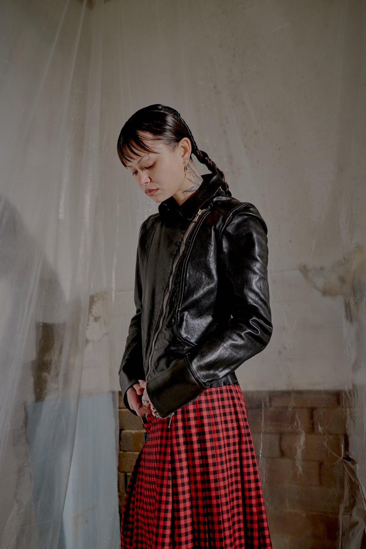 Person wearing a black leather jacket and red plaid skirt standing in a room with plastic sheeting.