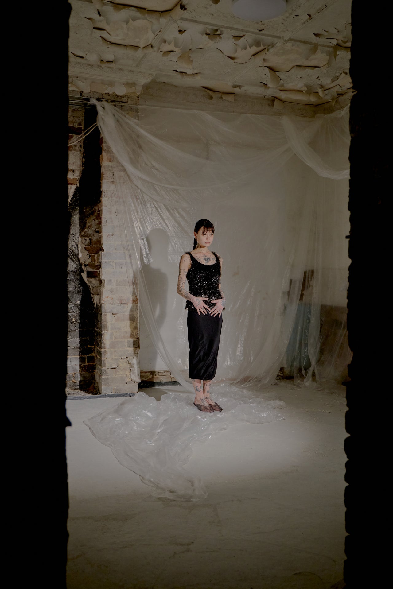 Person in a black dress standing in a room with white fabric and brick walls.