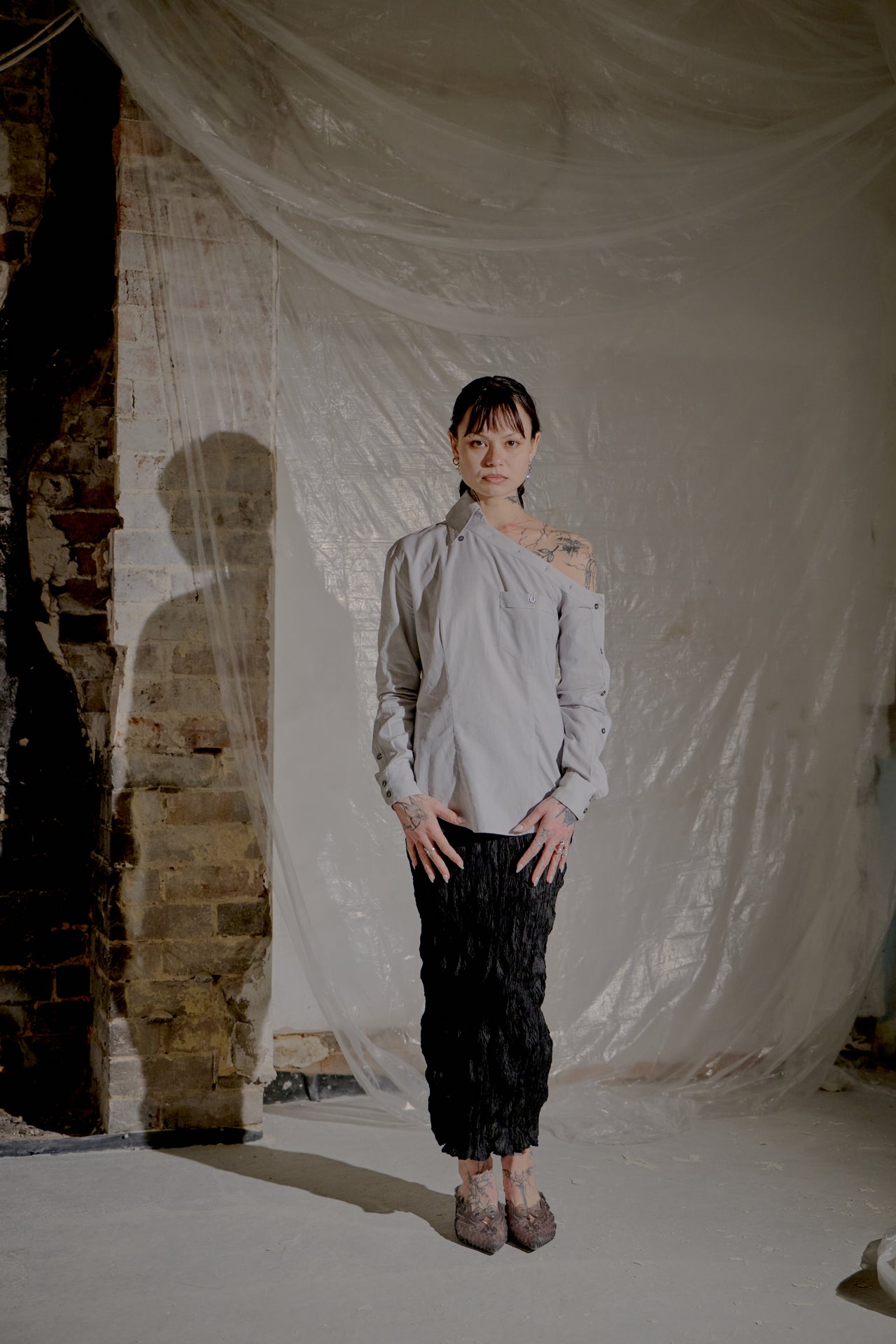 Person wearing a light grey Alexander McQueen top and black pants standing in front of a draped white fabric with a brick wall background.