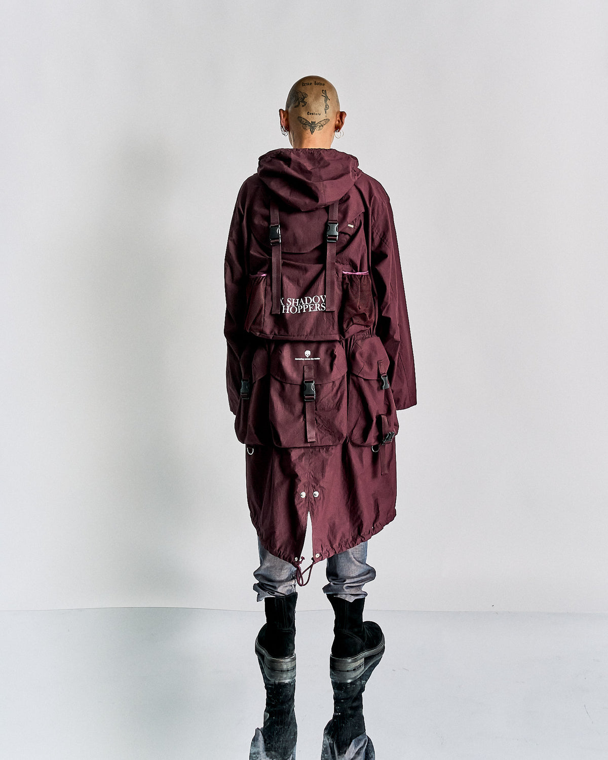 A model walks down the runway in a deep purple, Undercover layered outfit featuring a belted coat and cap, exuding a modern, edgy vibe. Dark boots complete the look.