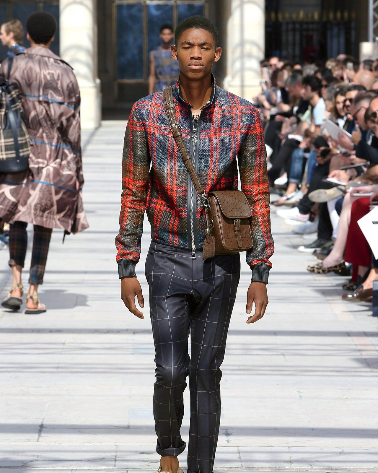 A male fashion model walks down an outdoor runway wearing a red and blue plaid Louis Vuitton jacket, checkered pants, a brown crossbody bag, and sandals, with an audience seated along the sides.
