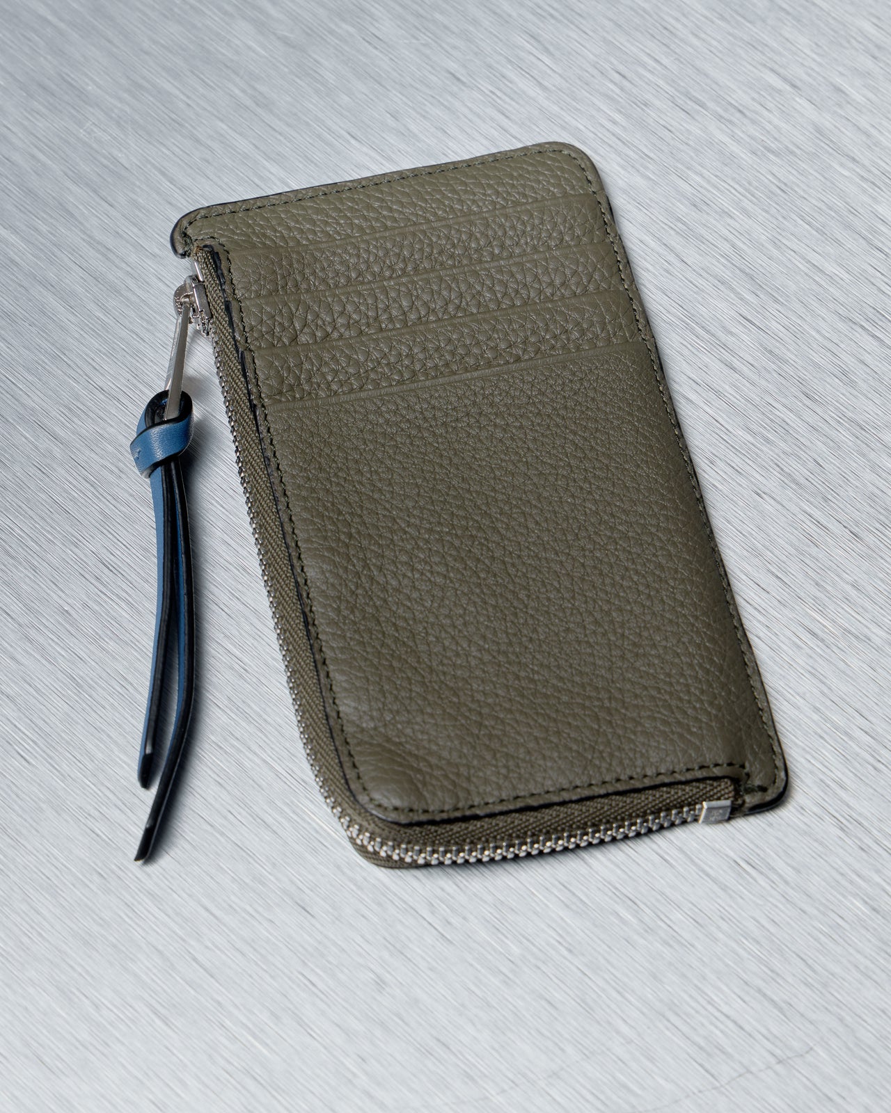 A textured Loewe leather wallet lies on a metal surface. The wallet has olive and brown sections with a zippered edge and a blue strap
