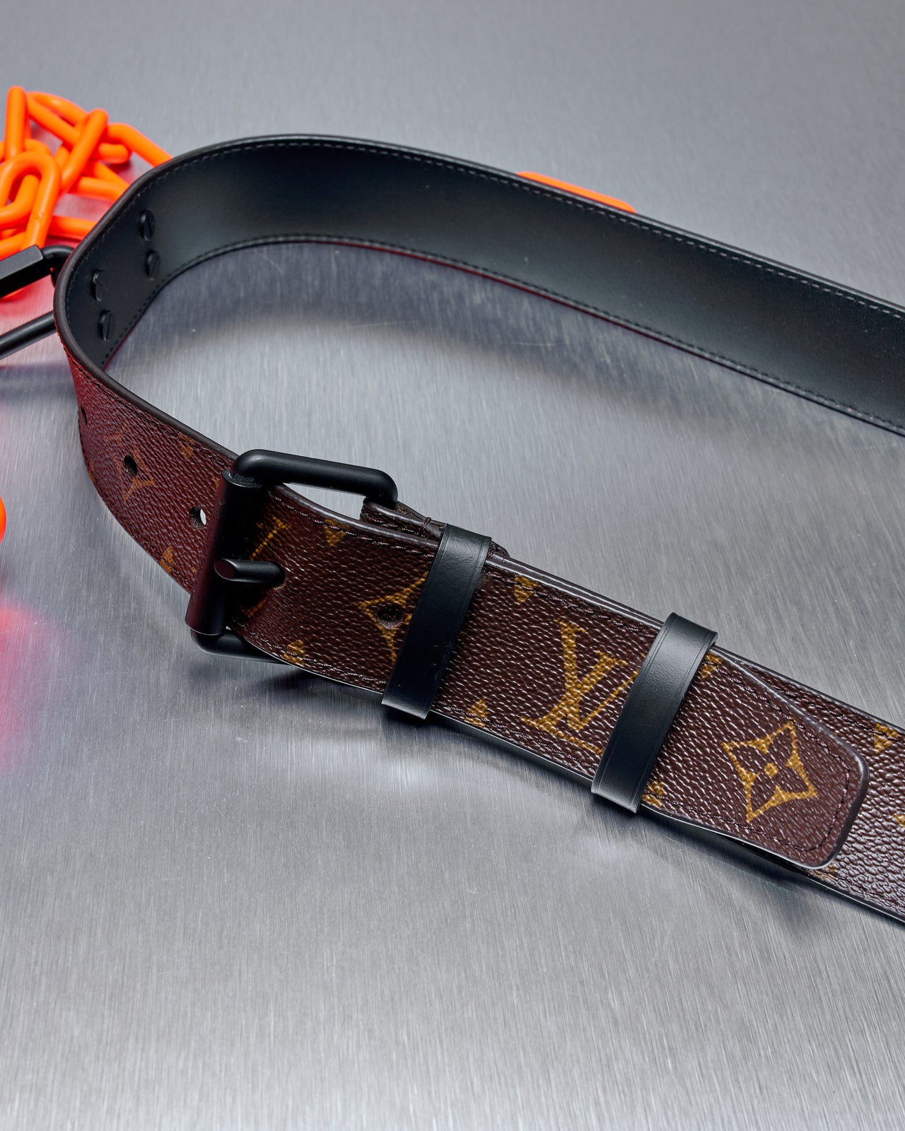 A Louis Vuitton belt with a monogram canvas and neon orange chain with detachable screw carabiner clips on a silver background.