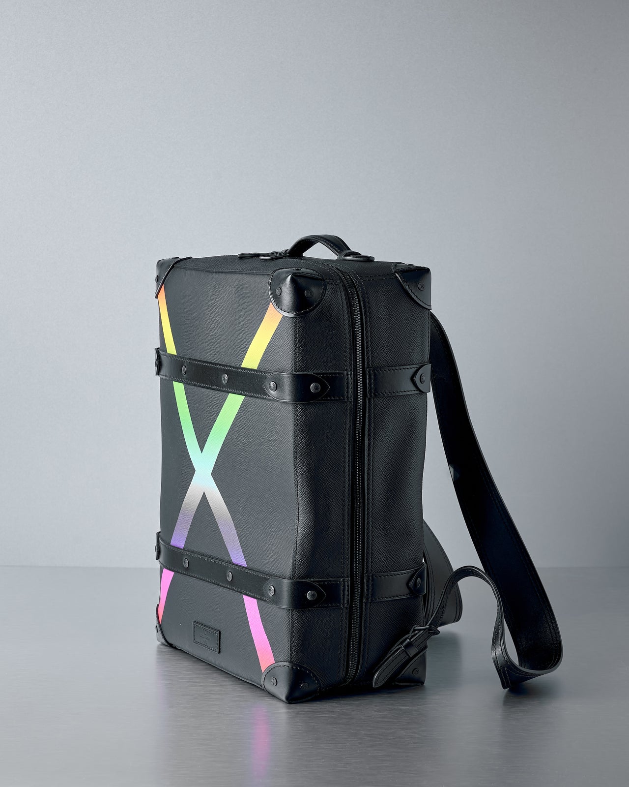 A sleek black Louis Vuitton suitcase with colourful rainbow gradient "X" on the front. It has black leather accents and a modern, stylish look, resting on a grey surface.