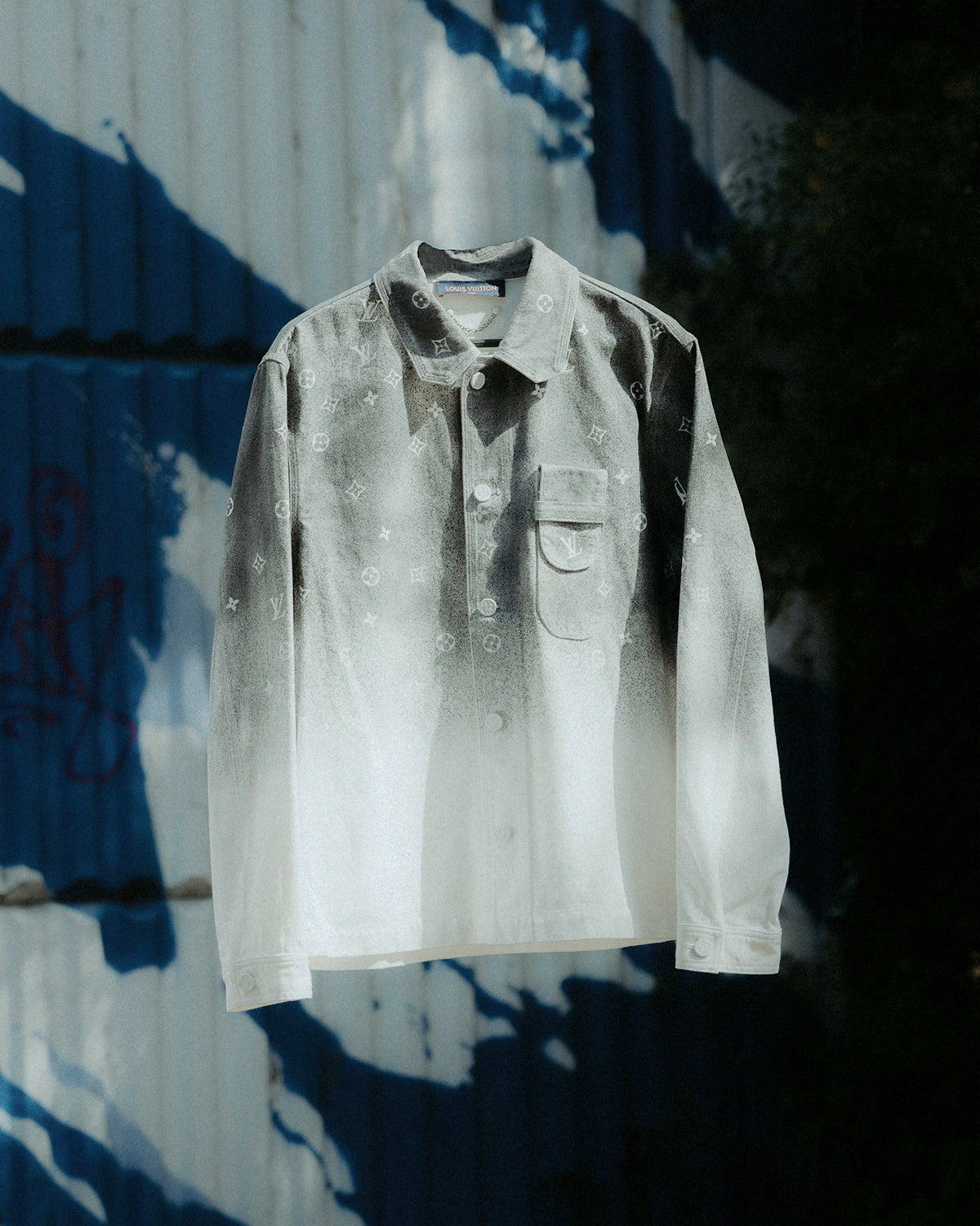 A shirt-style Louis Vuitton jacket with a gradient monogram denim overshirt, featuring a white and grey colour scheme and enamel button closures. It is displayed on a shadowy background with a blue wall in the background.
