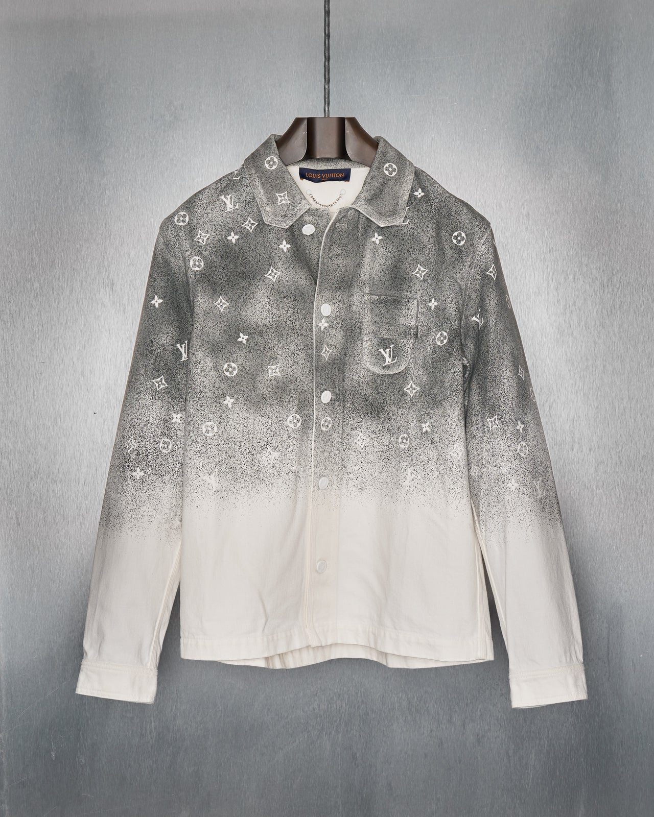 A shirt-style Louis Vuitton jacket with a gradient monogram denim overshirt, featuring a white and grey colour scheme and enamel button closures. It is displayed on a shadowy background with a blue wall in the background.