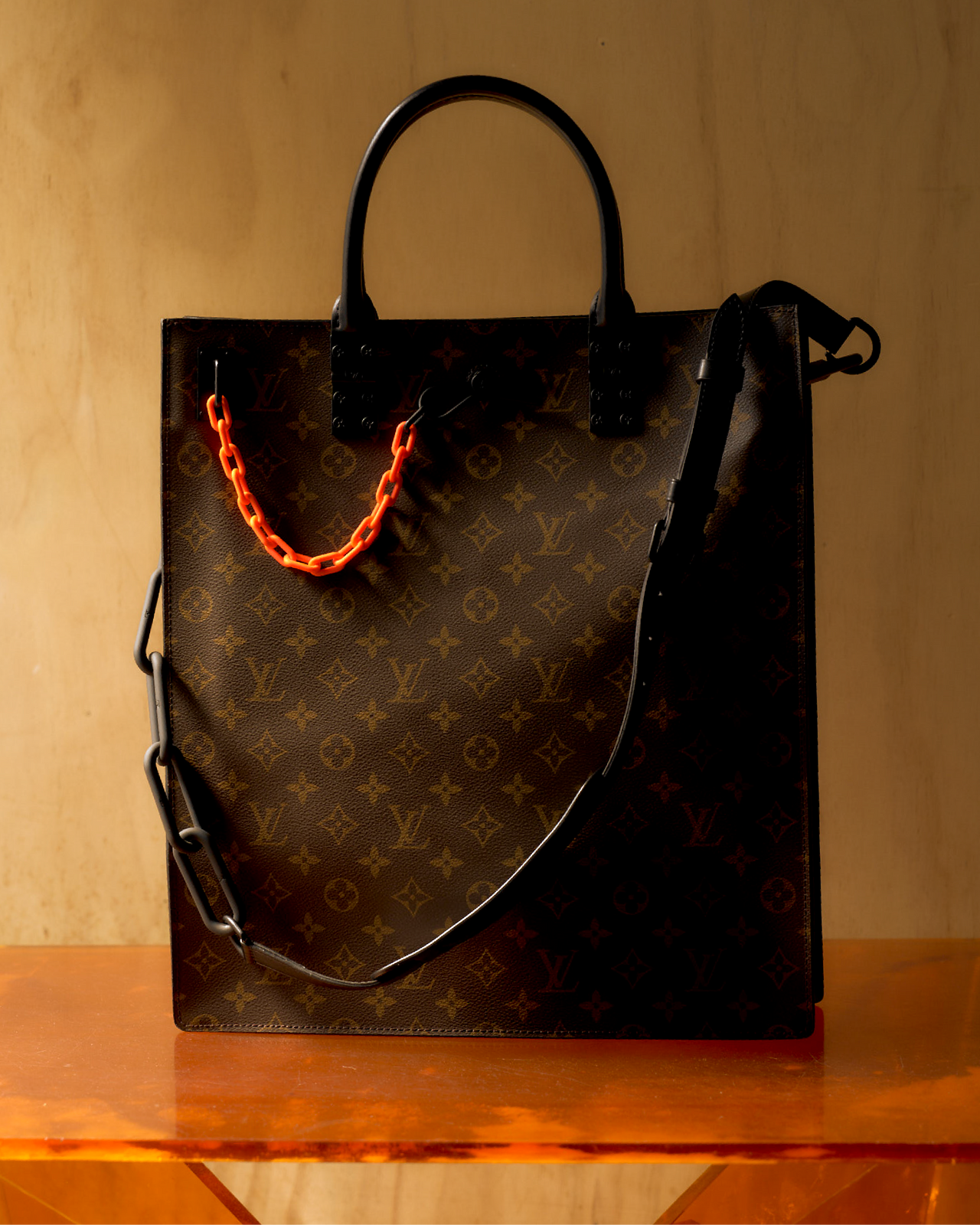 Louis Vuitton monogram tote bag with orange chain, matte black leather detailing, and a visible logo on the fabric.