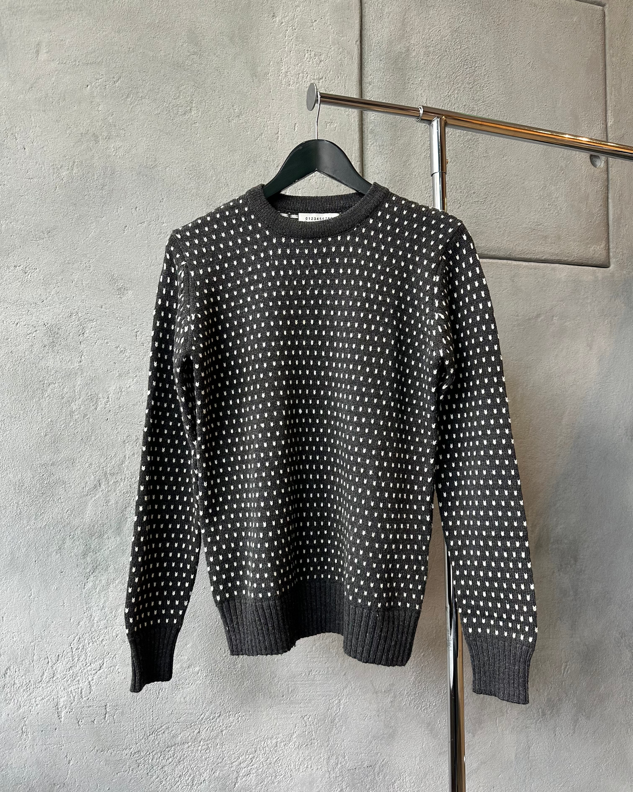 Charcoal Maison Margiela sweater with white pattern on a hanger against a gray wall