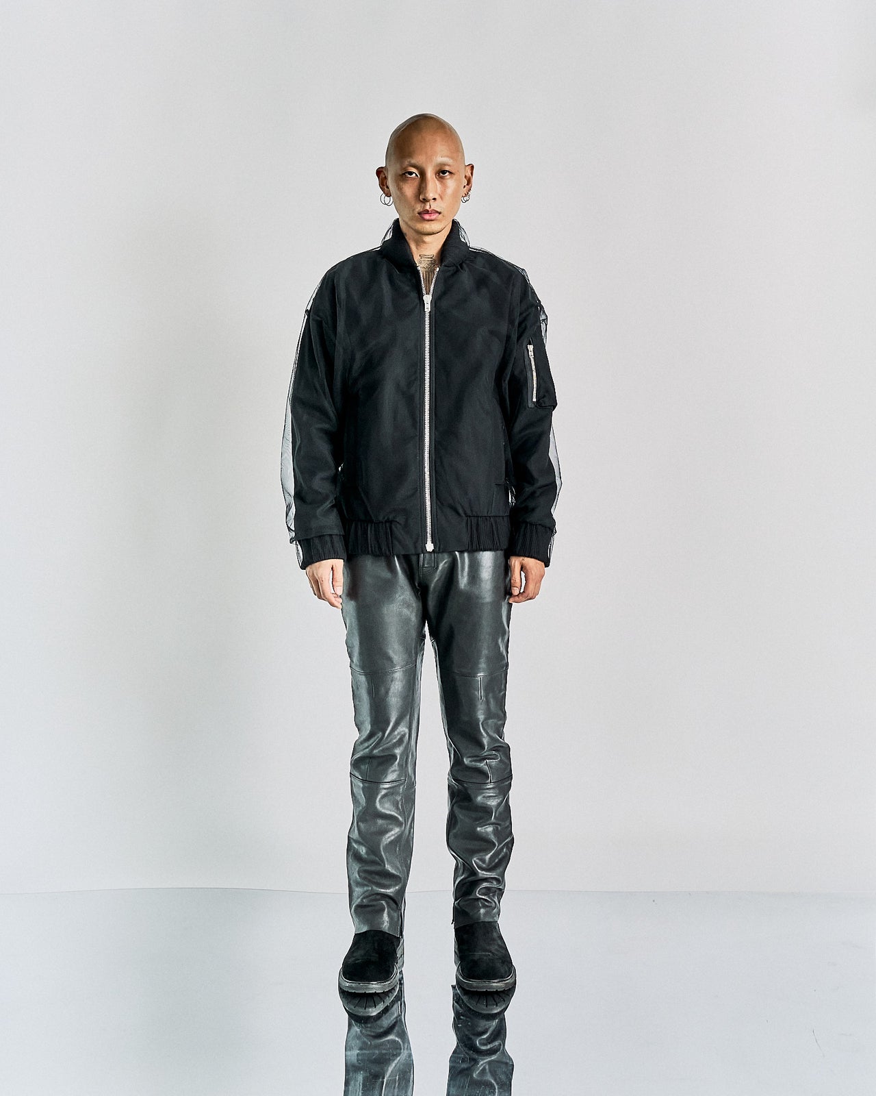 A model wearing a black Maison Margiela bomber jacket with a tulle overlay and ribbed finishings, paired with black leather pants. The model has a shaved head and is posing with one hand to the side.