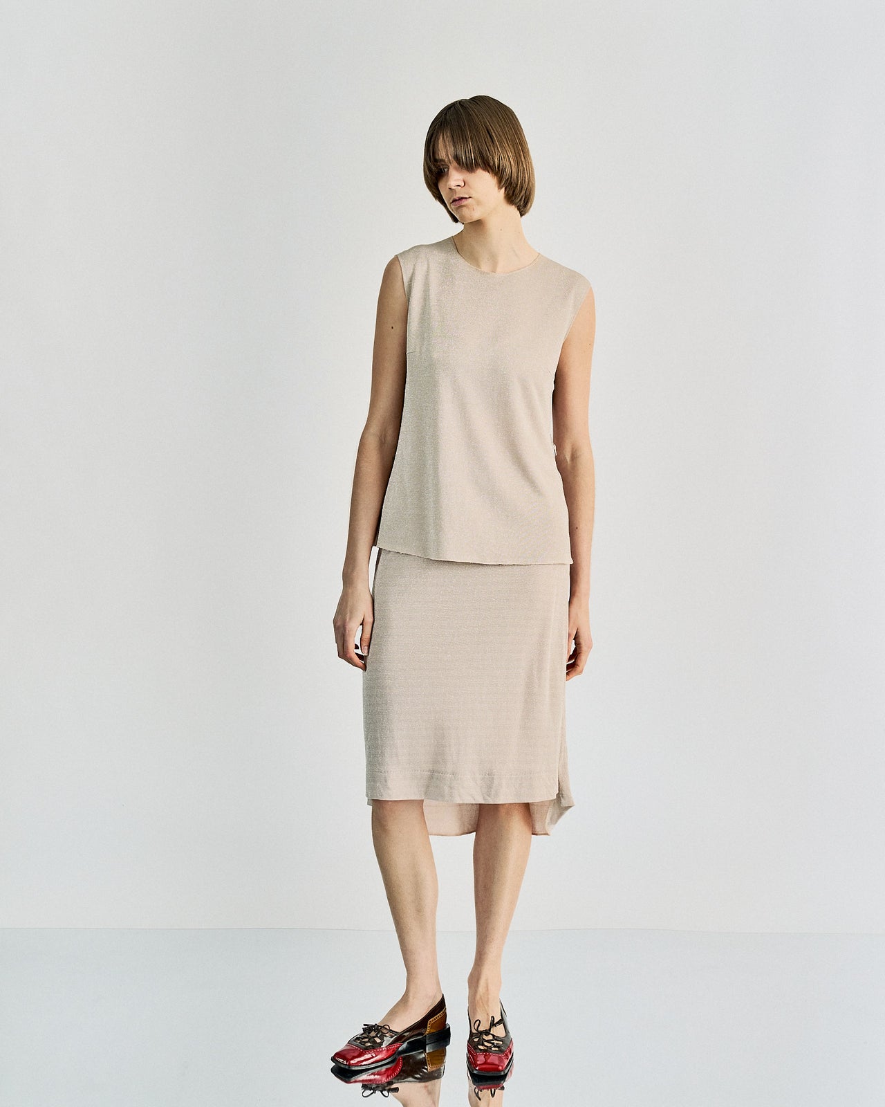 A person with short hair is wearing a sleeveless beige top and matching knee-length skirt from Maison Margiela , standing against a plain white background, looking peaceful.