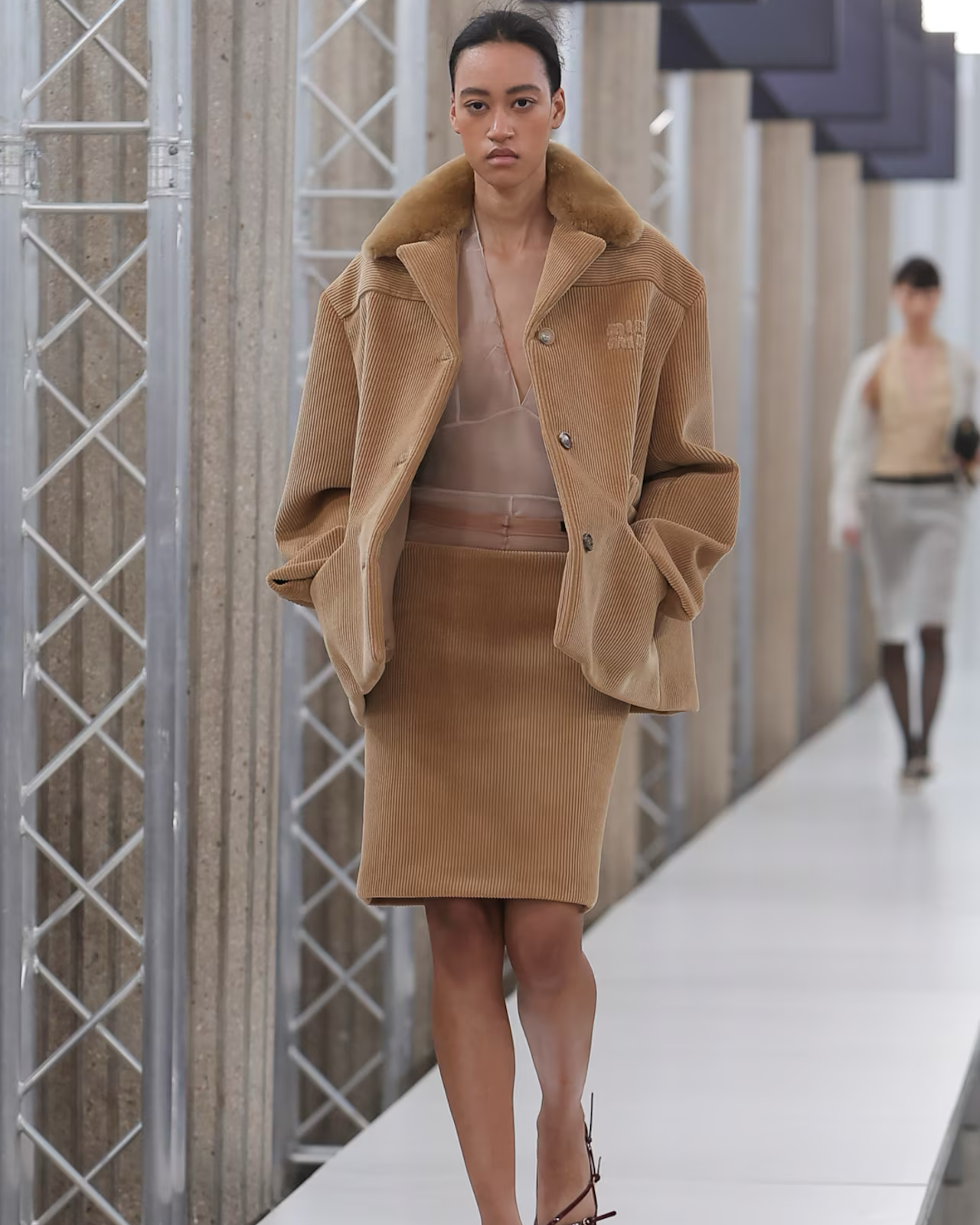 Model walking on a Miu Miu runway wearing a beige coat and skirt set.
