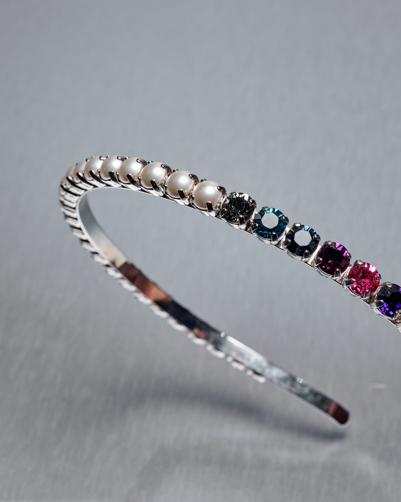 A silver Miu Miu headband adorned with pearls and colorful gemstones is shown on a gray background. The gems, in varied hues, add a vibrant touch to the elegant piece.