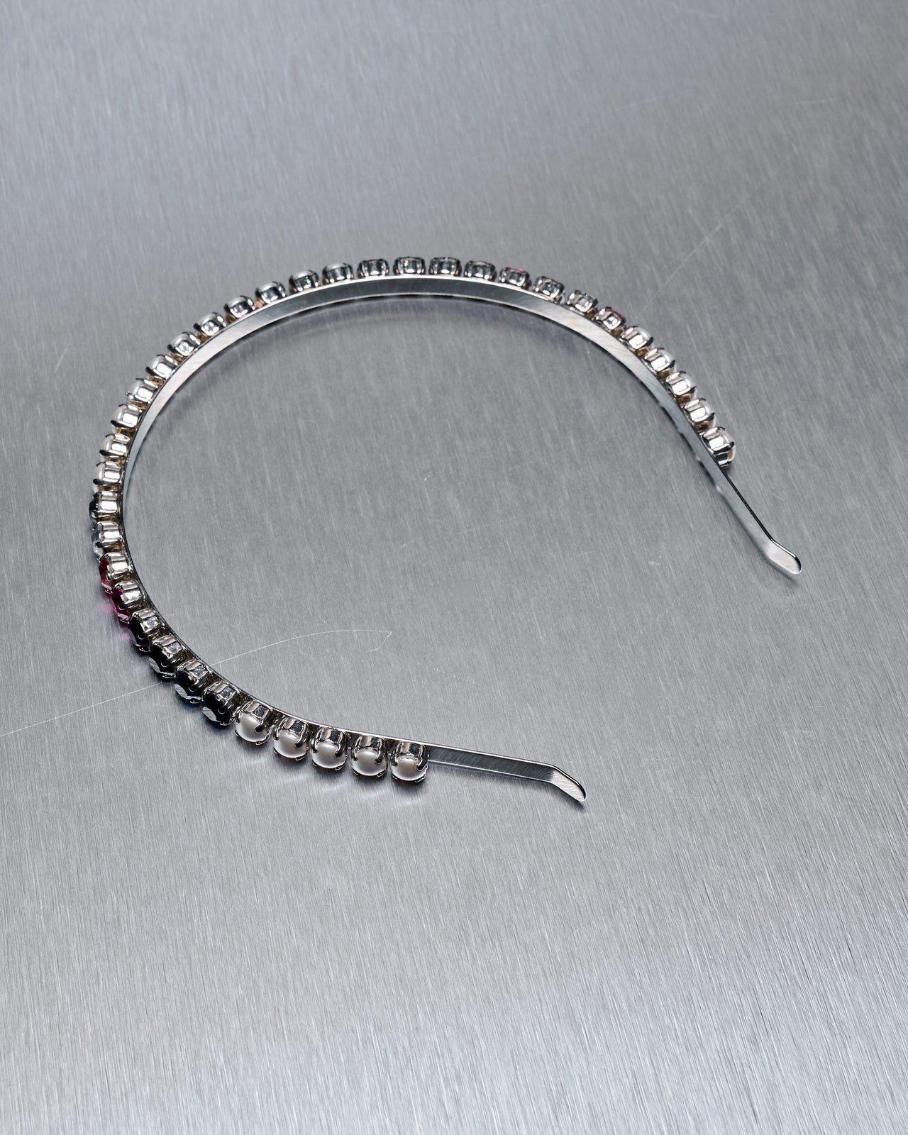 A silver Miu Miu headband adorned with pearls and colorful gemstones is shown on a gray background. The gems, in varied hues, add a vibrant touch to the elegant piece.