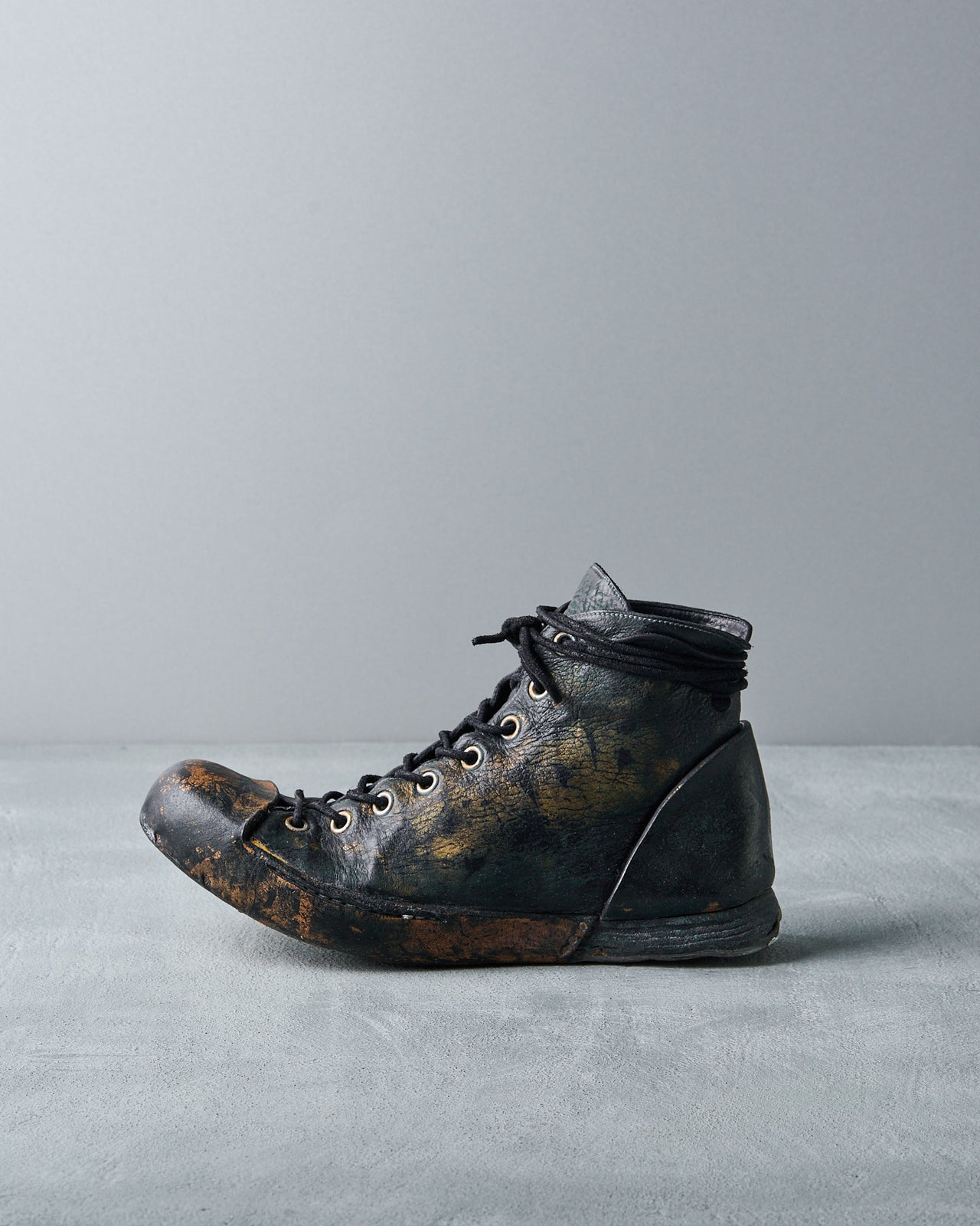 A Nihomano black boot with visible scuffs and creases. It features laces threaded through metal eyelets and sits on a plain grey background.