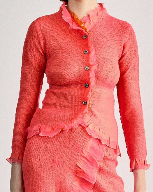 A woman wearing a pink ruffle Peplum by Yoshiki Hishinuma cardigan with black Mother of Pearl buttons and an orange pleated skirt with floral patterns. She is accessorized with a red tie and black boots.