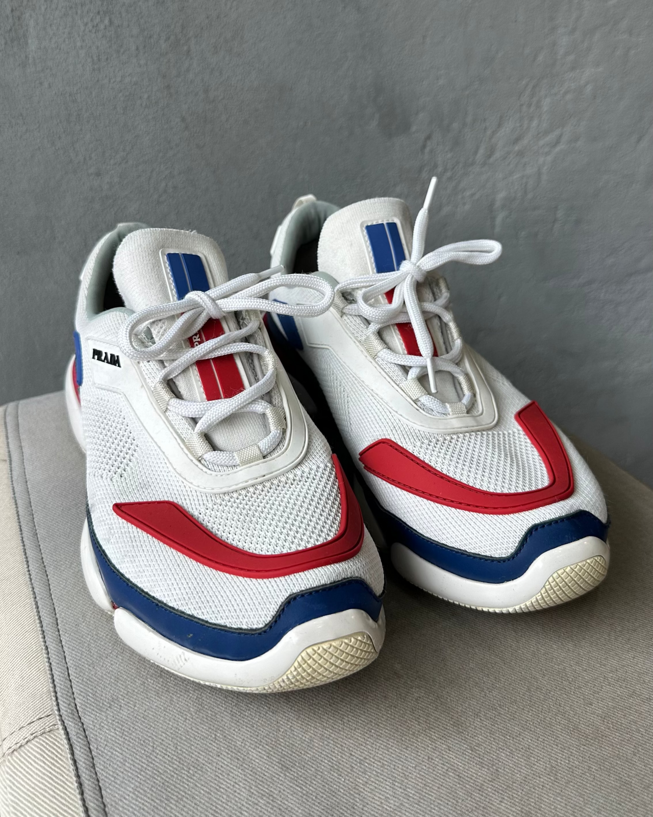 White Prada sneaker with blue and red accents on a gray surface against a gray background