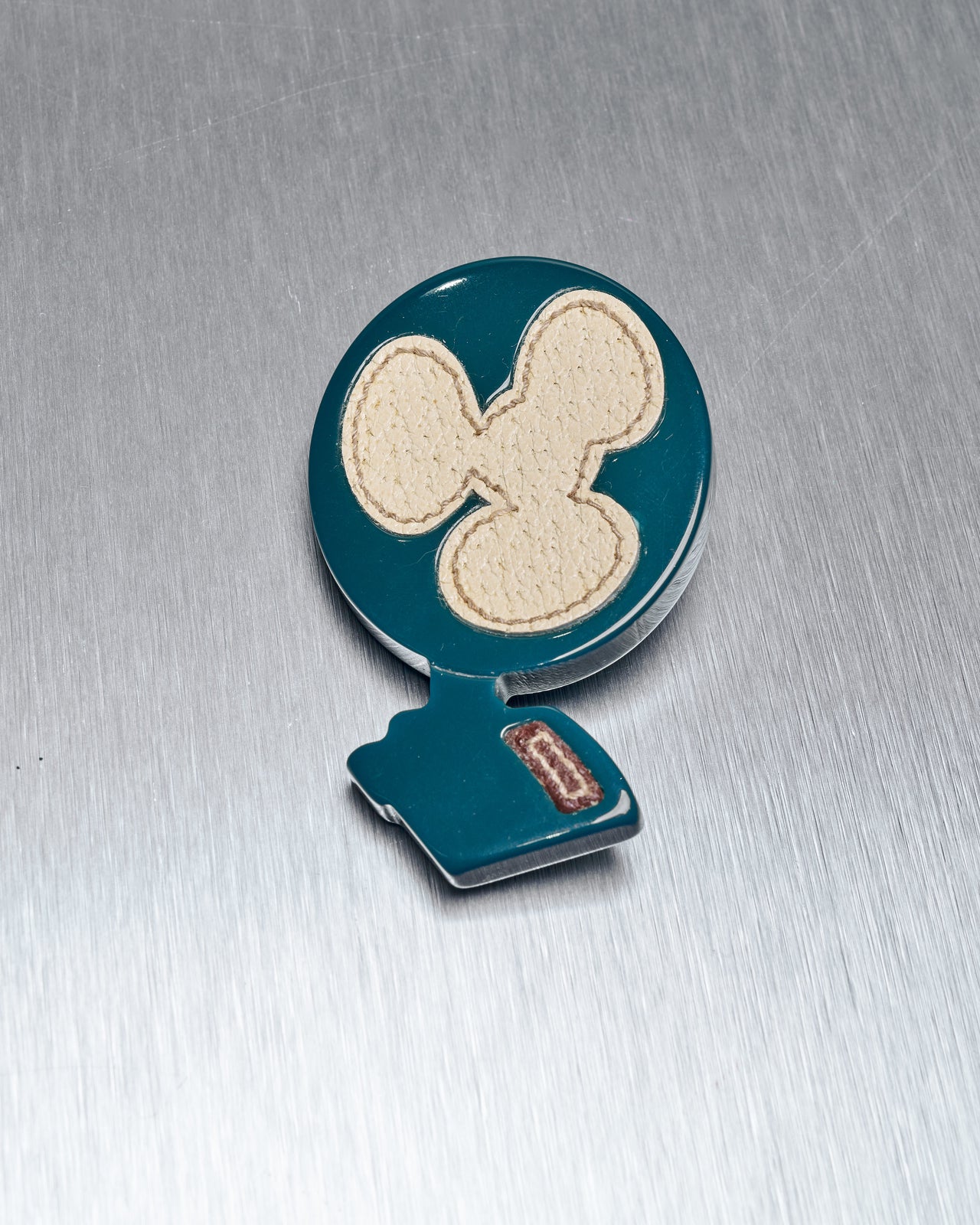 A Prada leather and enamel fan pin brooch with a blue background and acrylic fan-shaped design, featuring leather details, on a metallic surface.