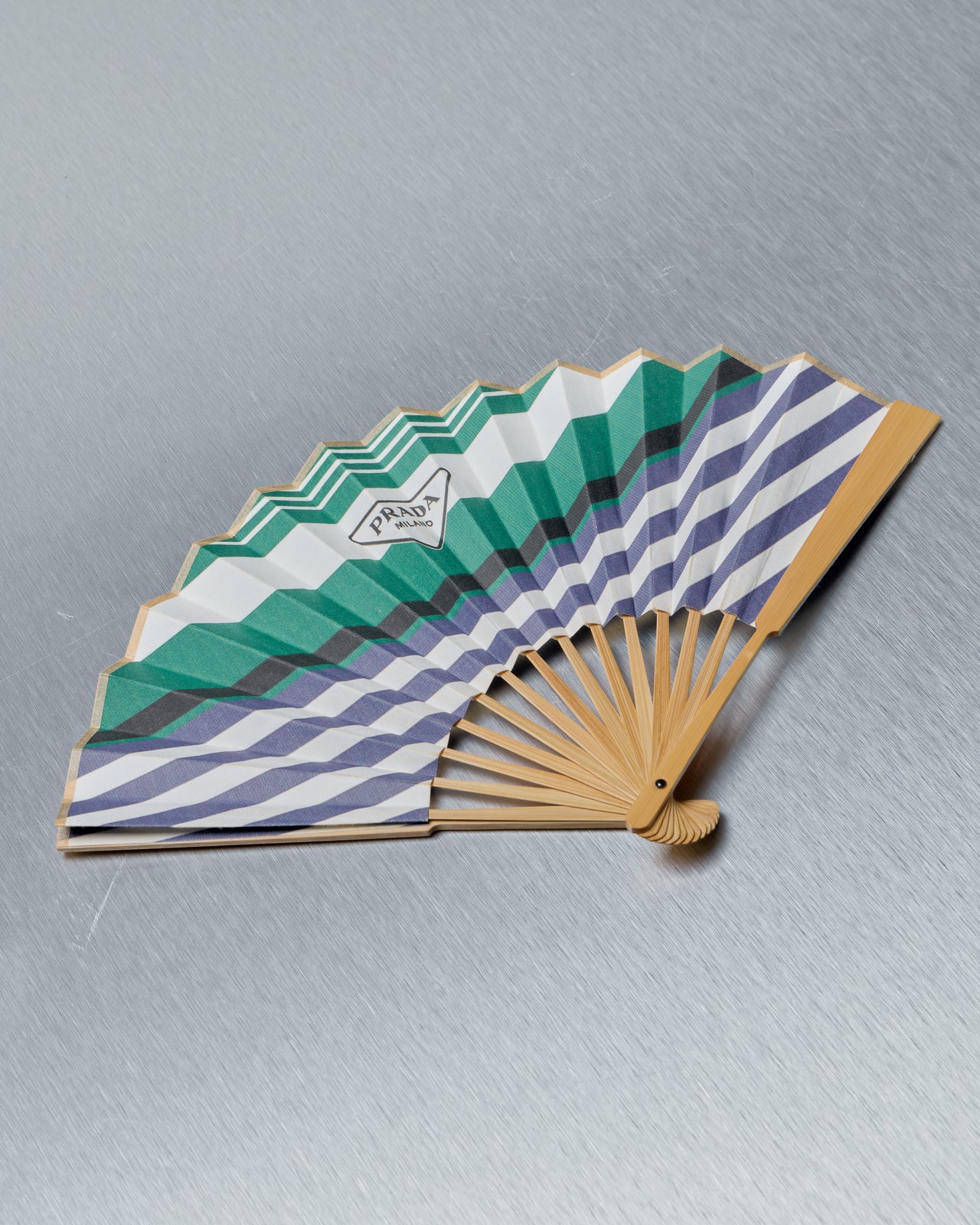Handheld Prada fan with green and blue striped design on a gray background