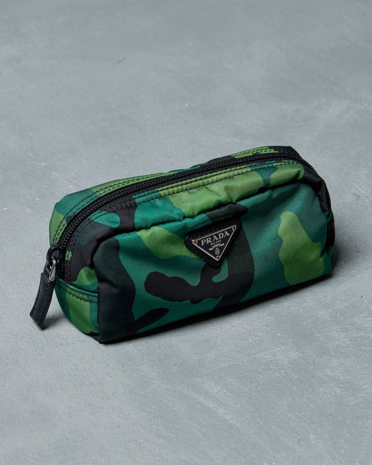 Green camouflage Prada pouch with black accents on a gray textured surface, featuring a prominent "Prada" logo on the front.