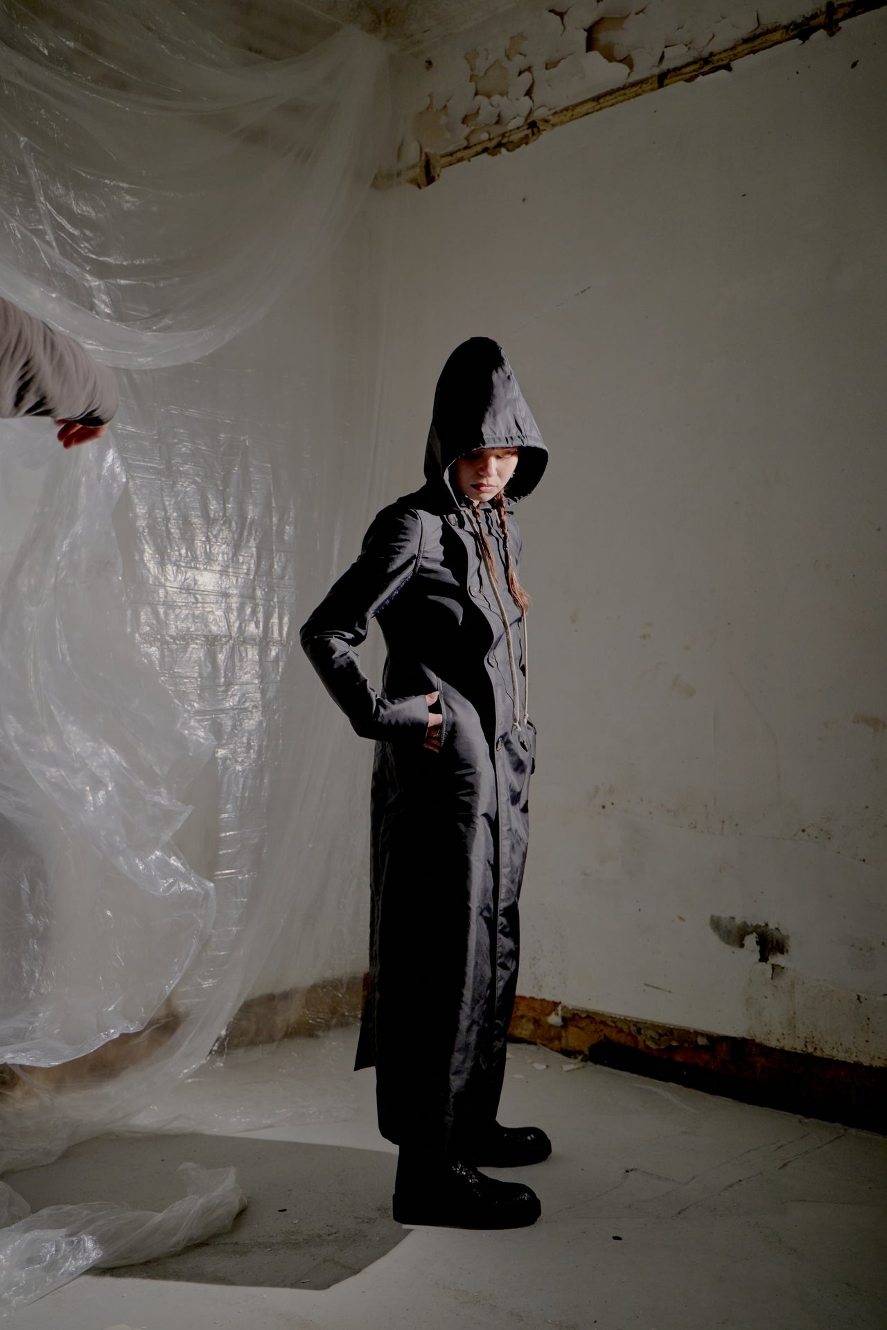 Person wearing a long black Rick Owens coat in a room with white walls and draped fabric.