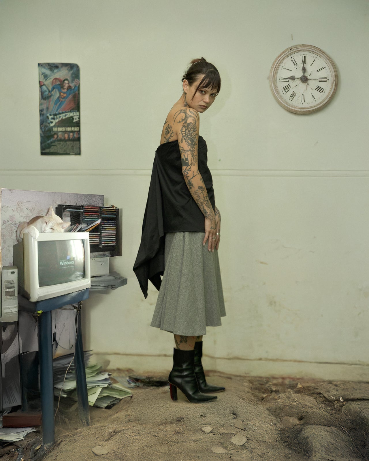 A person standing in a room with a computer monitor, wearing a Rick Owens bustier, a knee-length skirt, and boots. The room has a clock on the wall and appears to be cluttered.